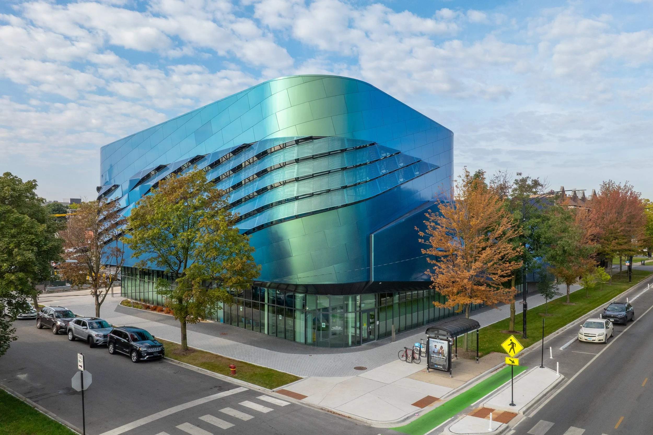 Modern building with a blue-green reflective facade, surrounded by trees with autumn foliage, parked cars, and a bus stop on a city street under a partly cloudy sky.