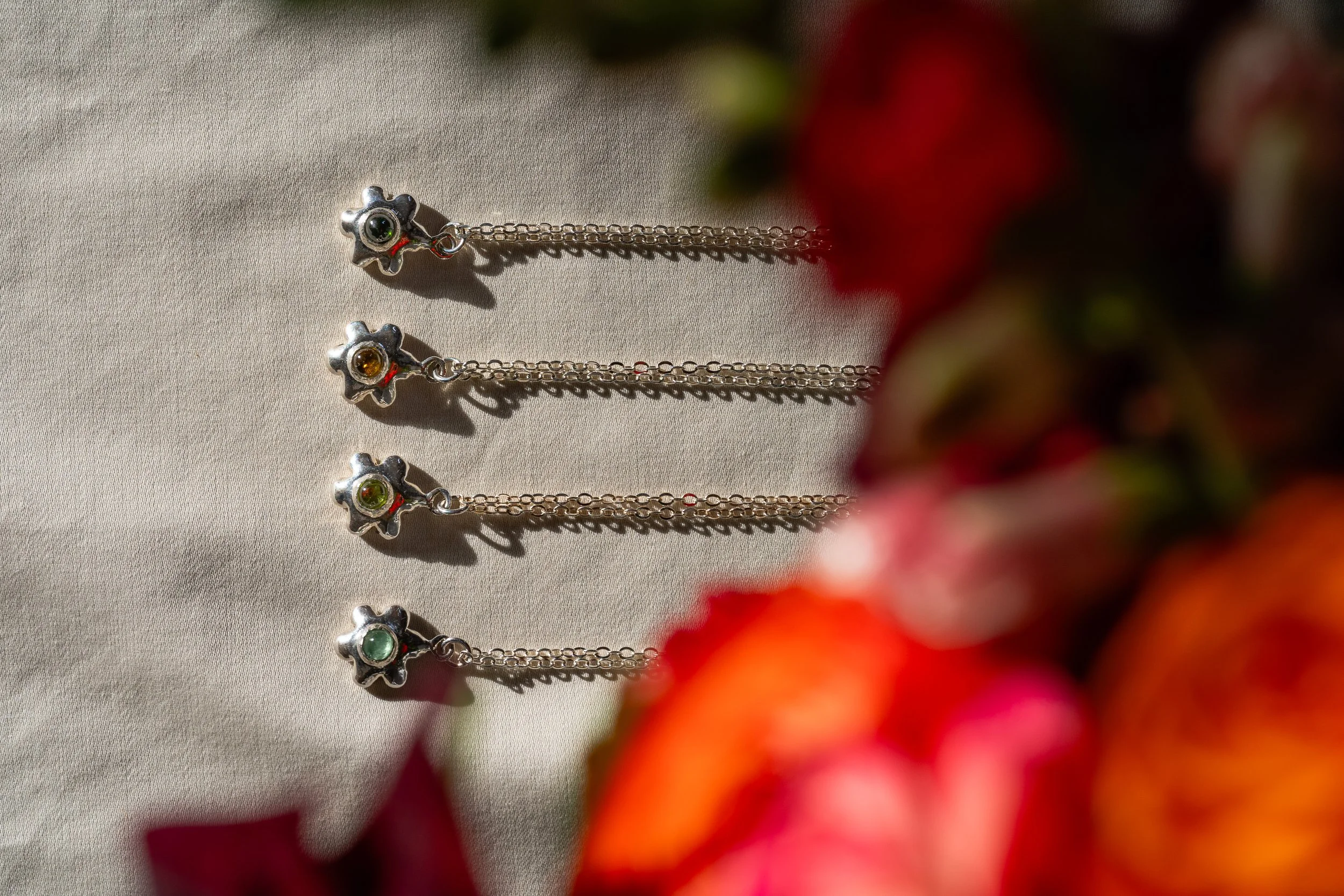 Four silver chain necklaces with star-shaped pendants, each containing different colored stones, laid out on a beige fabric surface with blurred red and pink flowers on the right side.