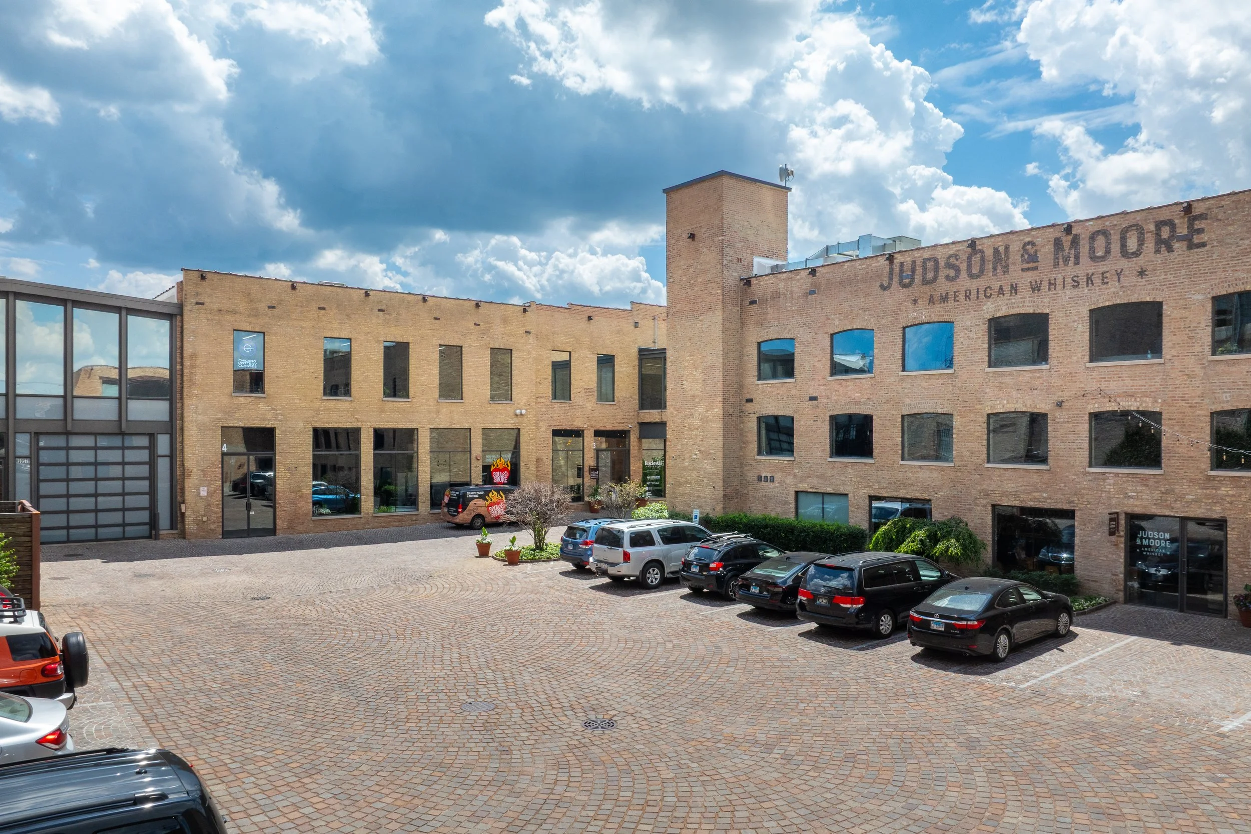 A parking lot in front of a brick office building with the sign Judson & Moore American Whiskey, and a partly cloudy sky overhead.