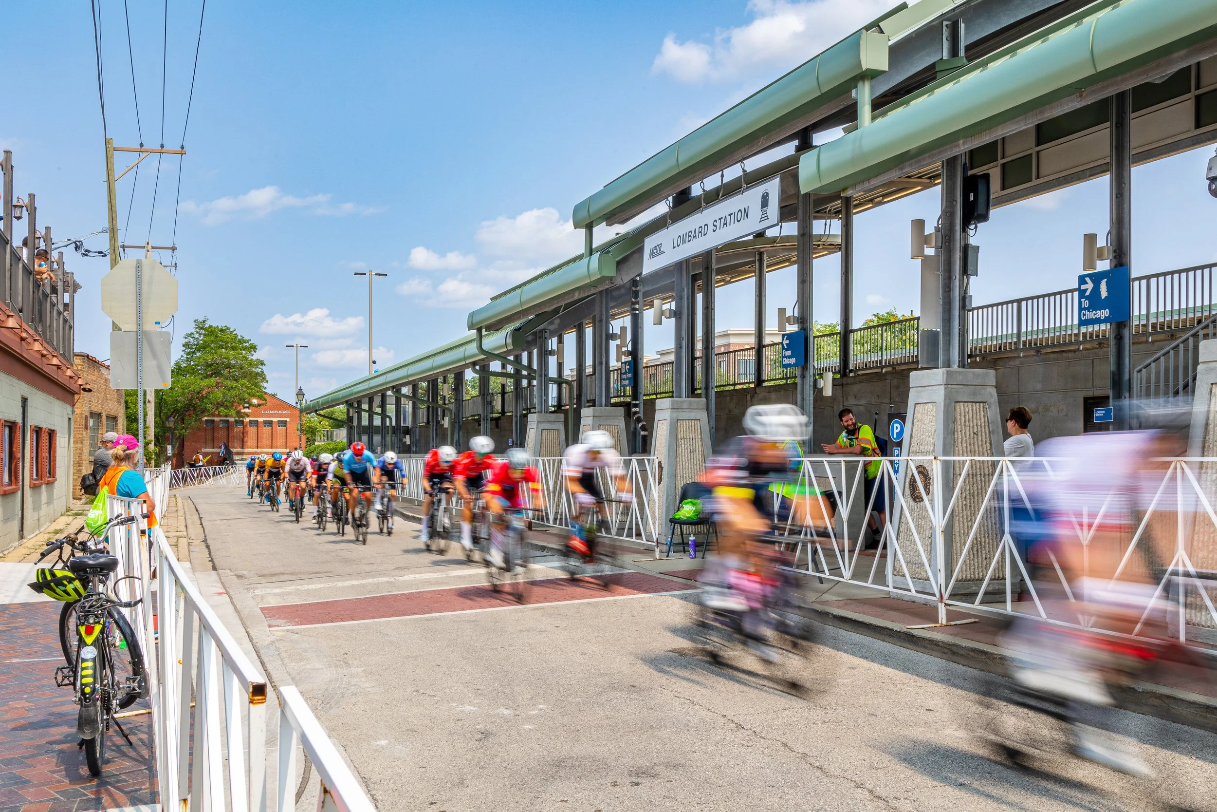 Cyclists racing near Lombard Station entrance, with spectators watching and barriers set up. Blurred motion of cyclists in the race, blue sky with clouds above.