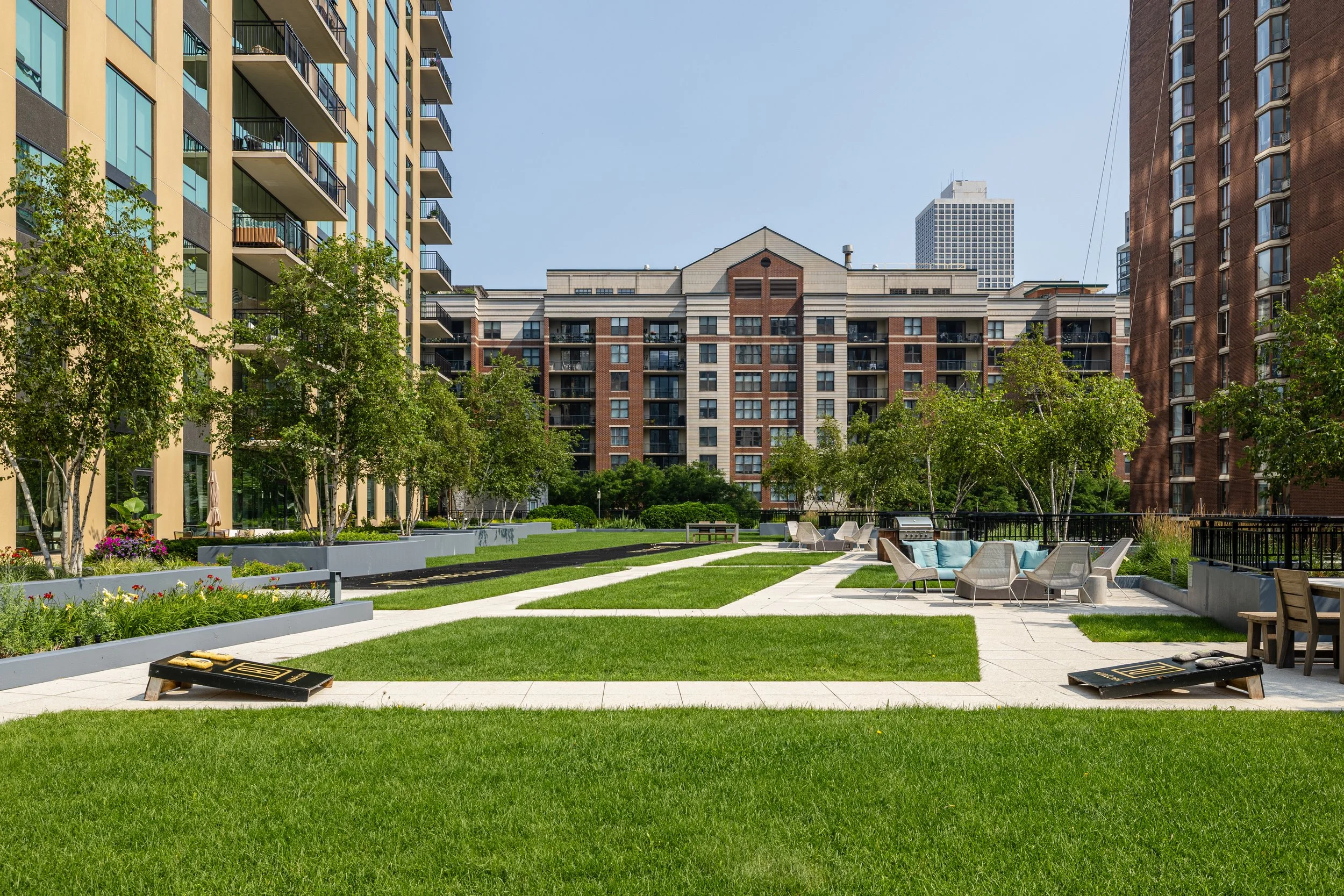 Outdoor courtyard with green grass, white pathways, and seating areas in front of tall residential buildings under a clear sky.