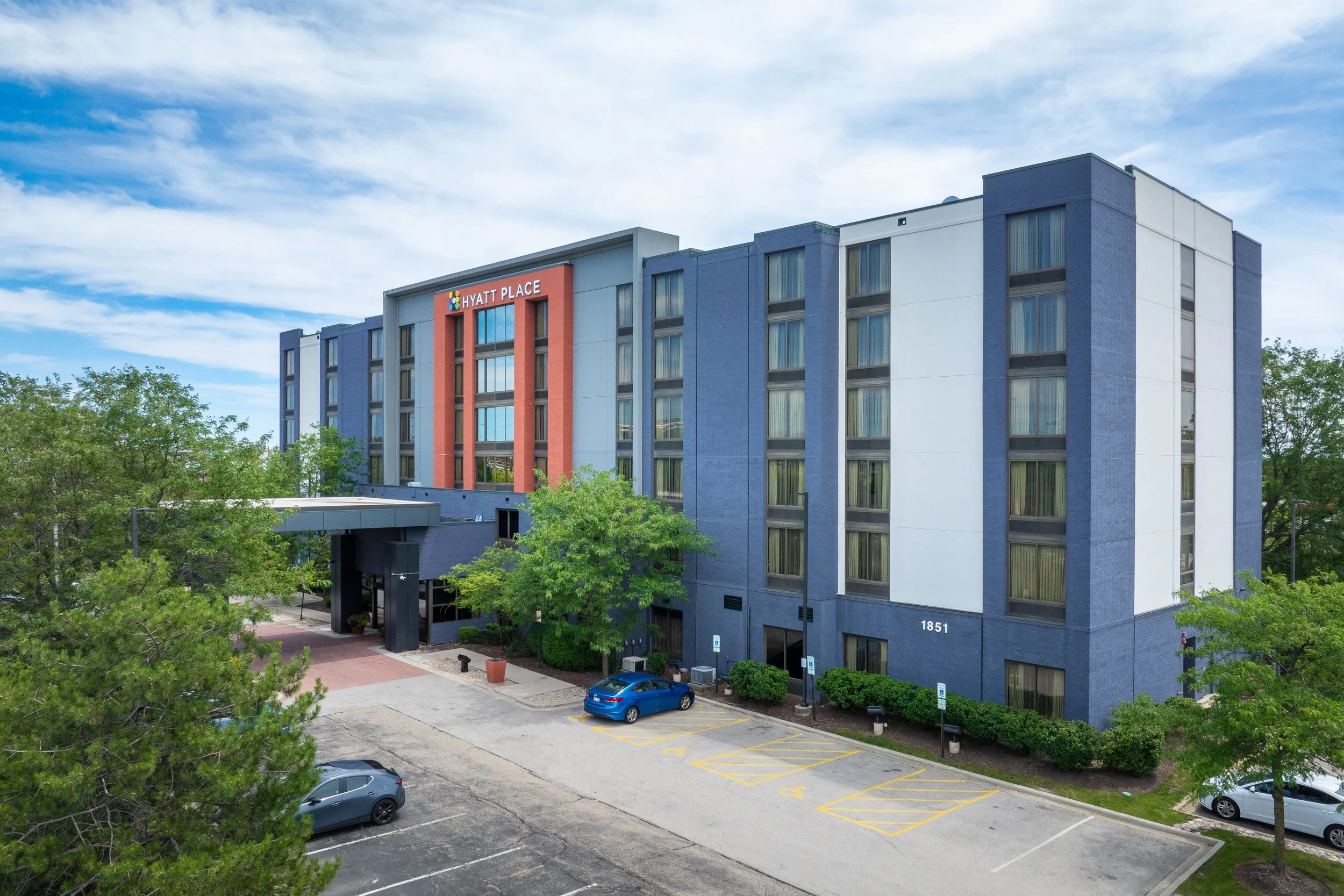 Photo of Hyatt Place hotel with colorful exterior, trees, parking lot with several parked cars, and a blue sky with clouds.