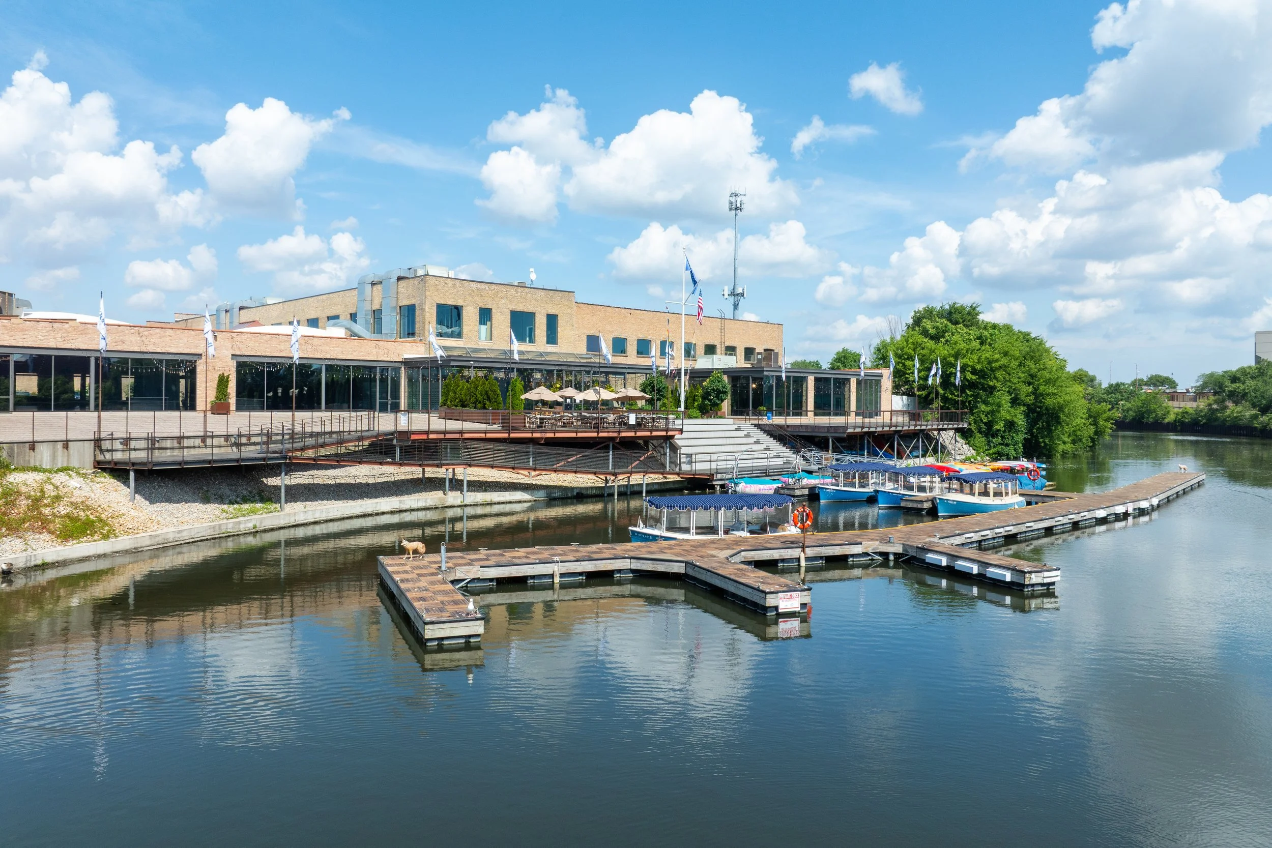 Buildings by a river with wooden docks and boats, outdoor seating area with umbrellas, lush green trees, cloudy blue sky.