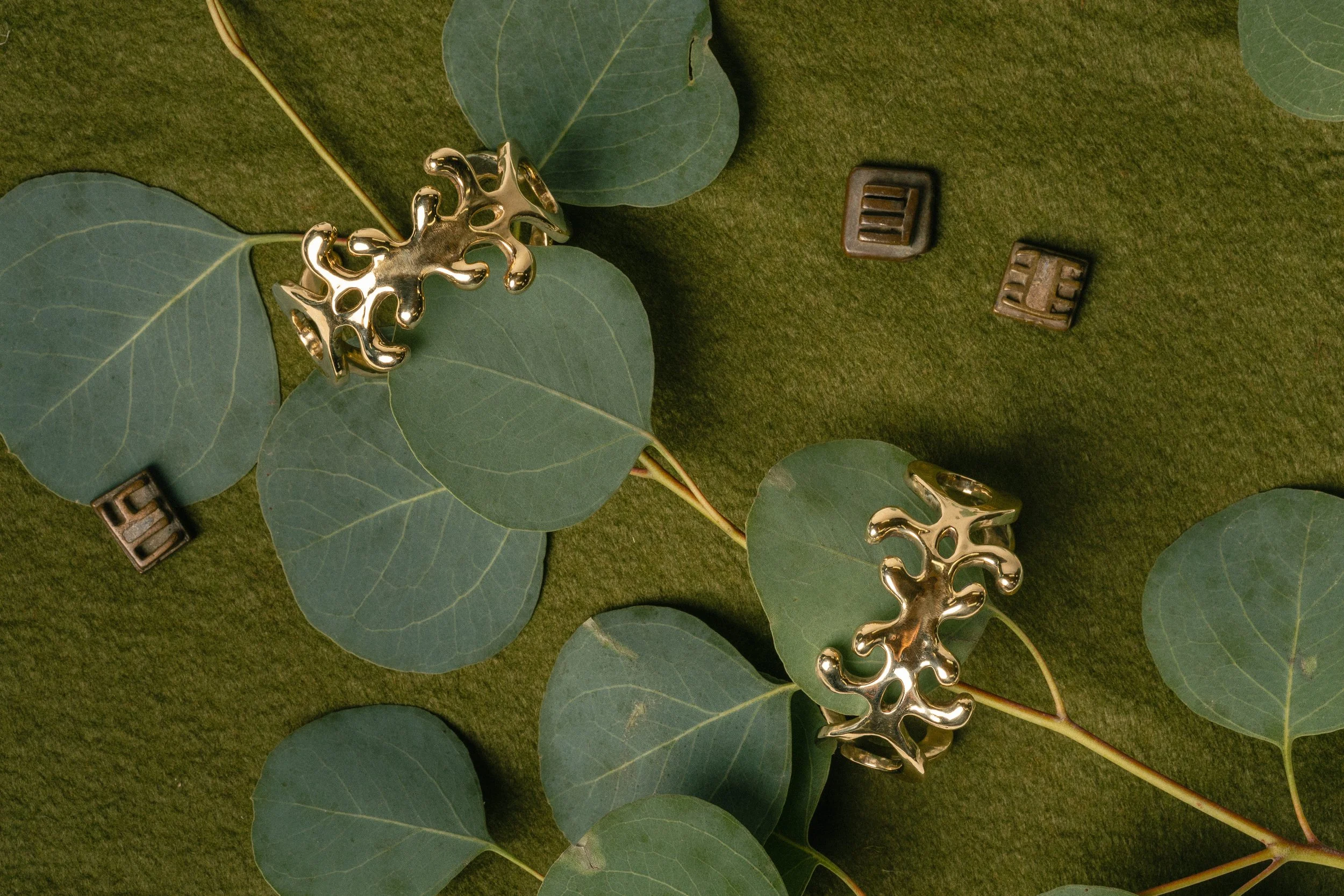 Gold treble clef earrings placed on green eucalyptus leaves and surrounded by small metal letter tiles on a green textured surface.
