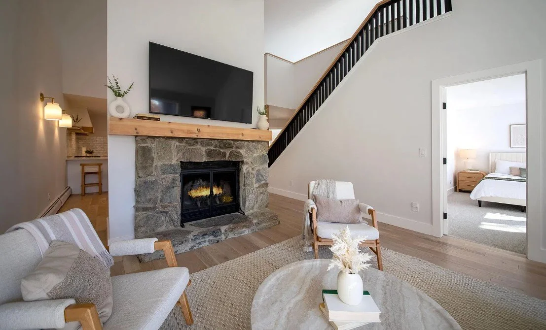 Living room with stone fireplace, wall-mounted TV, wooden staircase, and two chairs around a marble coffee table, with a bedroom visible through an open door.