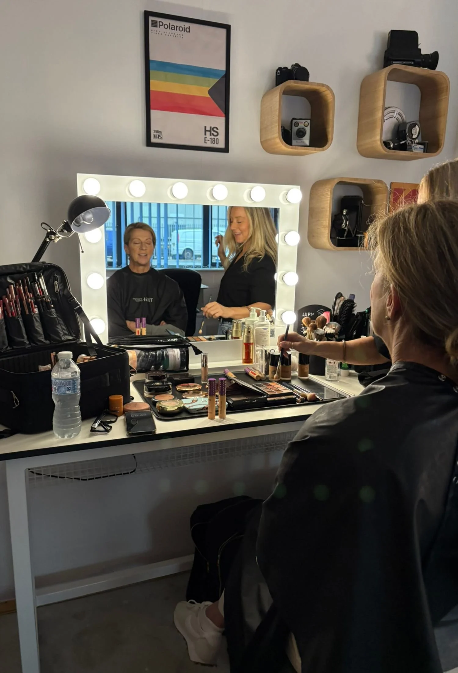 Makeup artist applying makeup on a woman sitting at a vanity mirror surrounded by makeup products, brushes, and beauty tools.