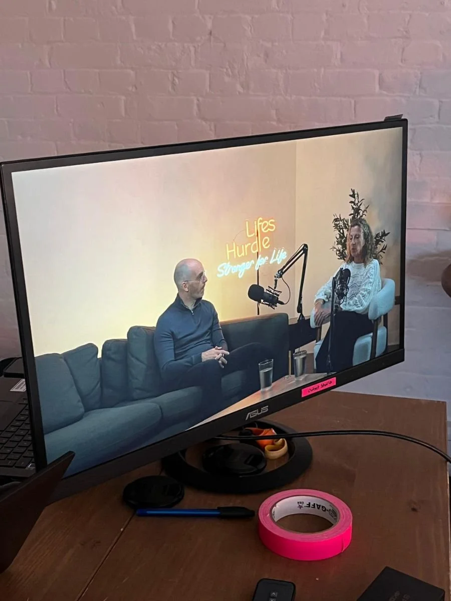 A computer monitor displaying a podcast recording session with two people, a man and a woman, sitting on a black couch in a studio with a yellow wall and a neon sign that reads "Lifes Hurdle Storage for Life." There is a microphone in front of each p