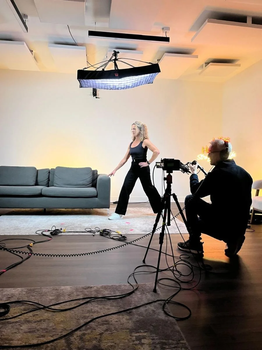 A woman stands on a film set in front of a gray couch with her hand on the armrest, while a cameraman kneels behind a camera on a tripod capturing her shot. The set features studio lighting, a modern ceiling, and a warm-toned background with a neon s