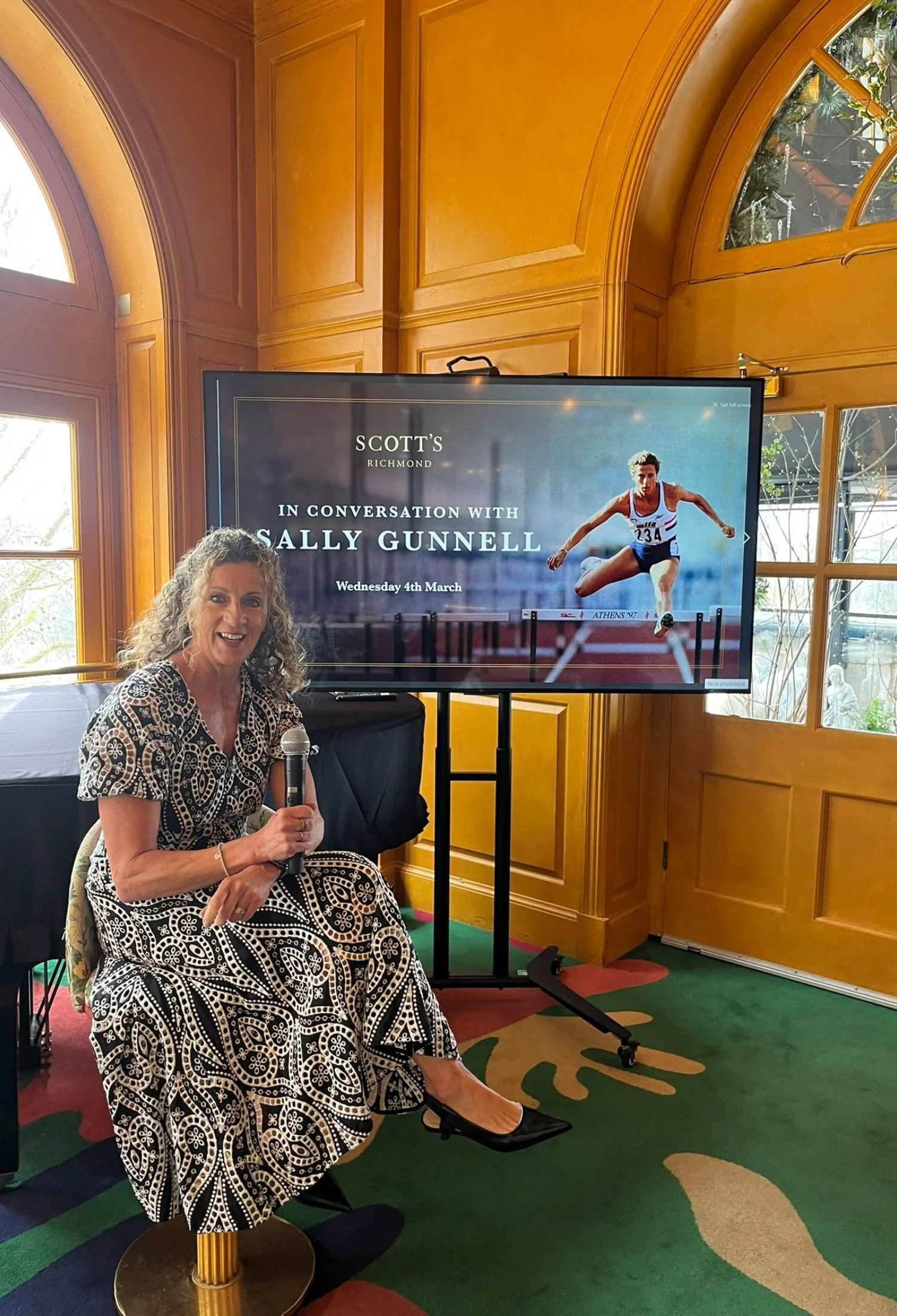 Sally Gunnell wearing a black and white patterned dress, sitting on a stool with a microphone in her hand, smiling at a presentation event. Behind her is a large screen
