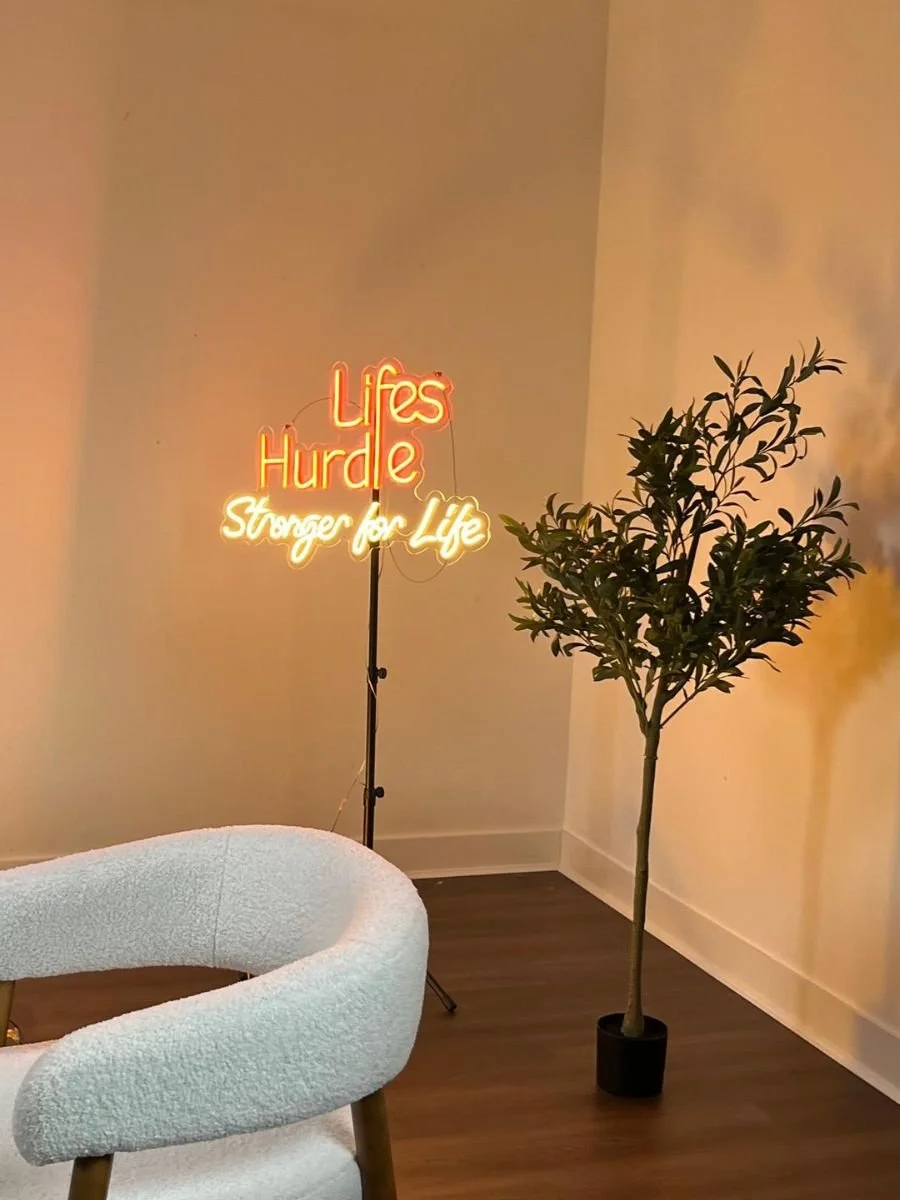 Interior with a neon sign reading 'Lifes Hurdle' and 'Stronger for Life', a potted plant with green leaves, and part of a white textured chair.
