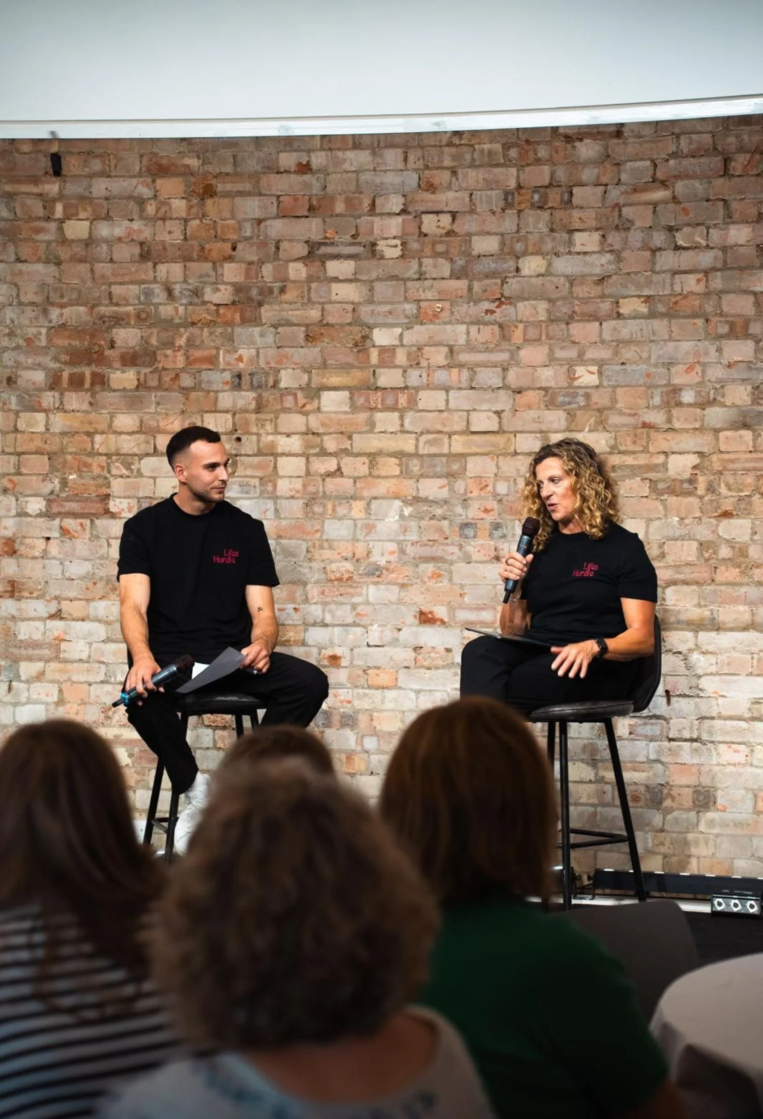Sally Gunnell sitting on stools in front of a brick wall, having a conversation while holding microphones during a panel discussion or interview, with an audience watching.