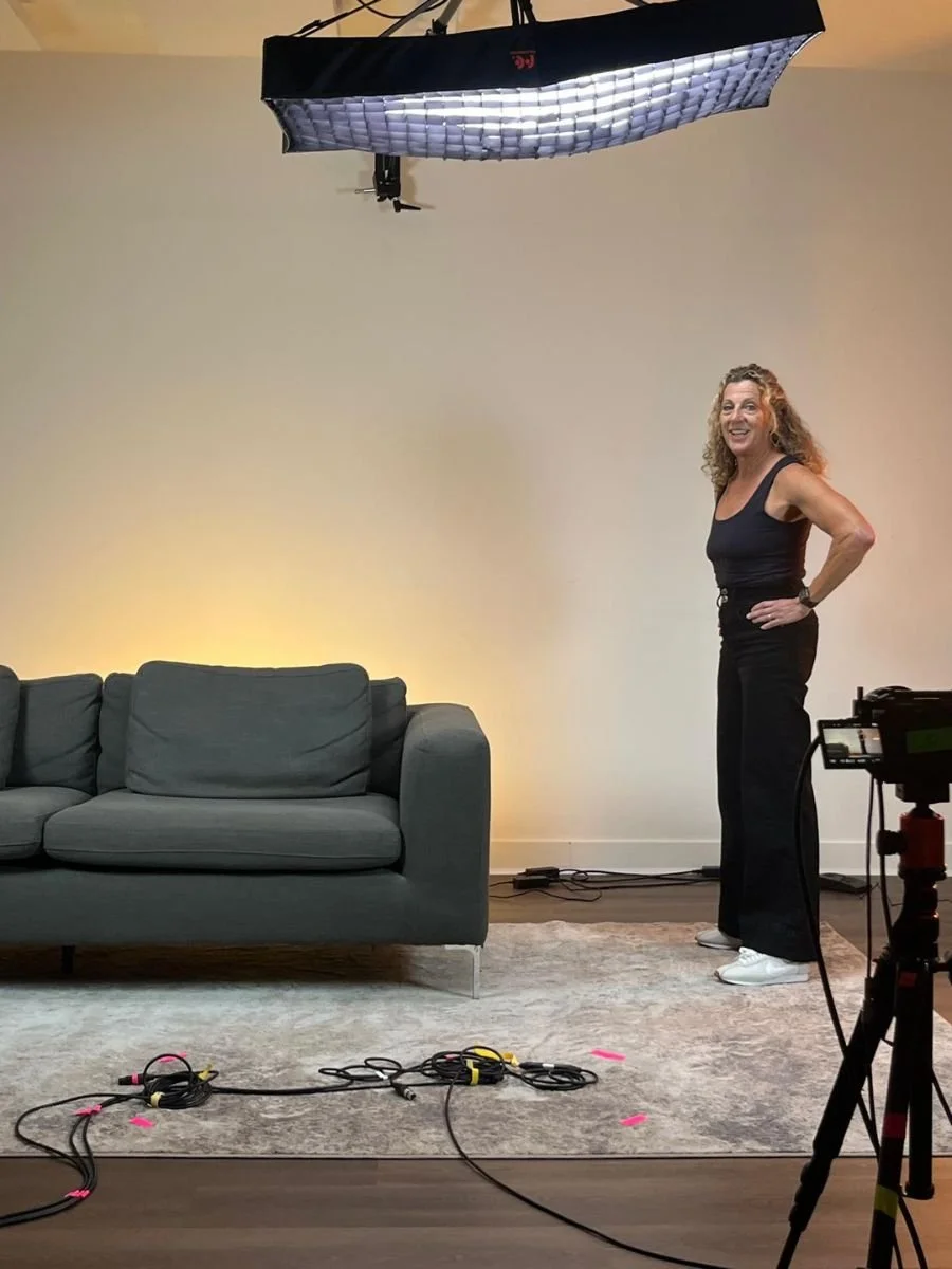 A woman with curly hair standing in a photography studio or set, smiling, next to a gray couch, with film and lighting equipment around, and a soft light on the wall behind her.