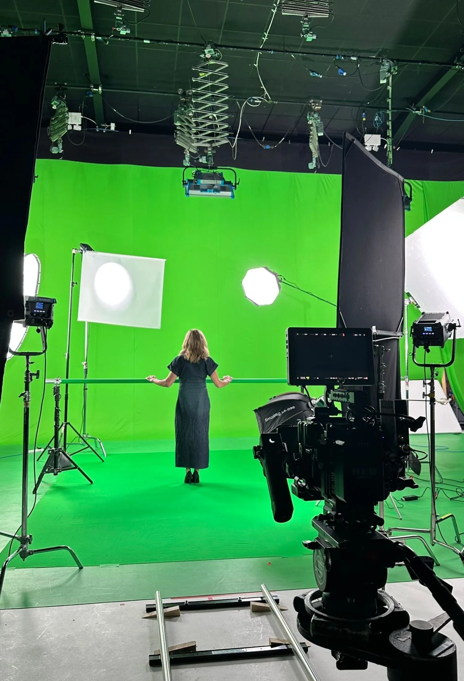A film set with a woman standing in front of a large green screen, surrounded by studio lighting and equipment.