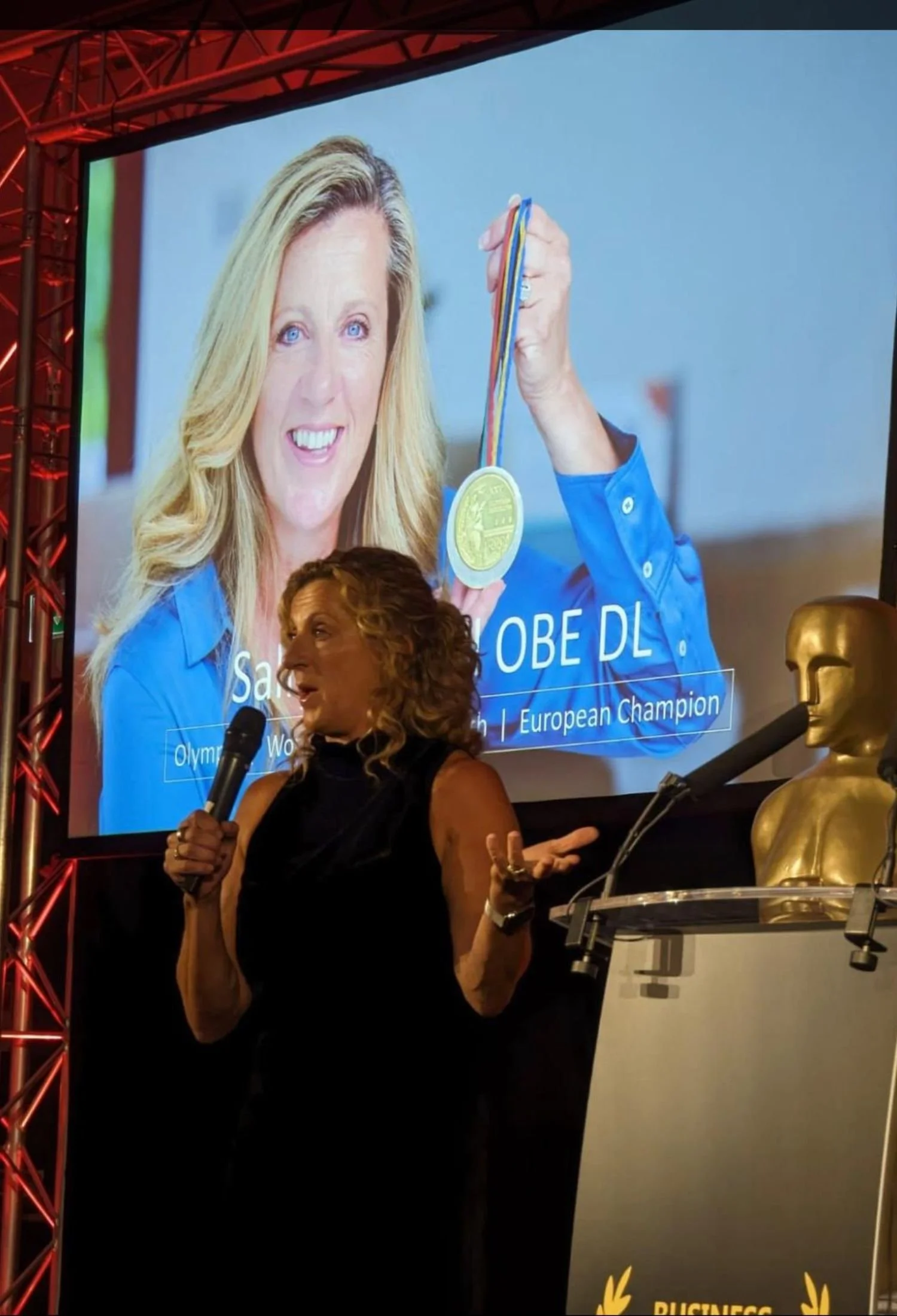 Sally Gunnell giving a speech at a podium , in front of a large screen displaying Sally Gunnell holding a gold medal.