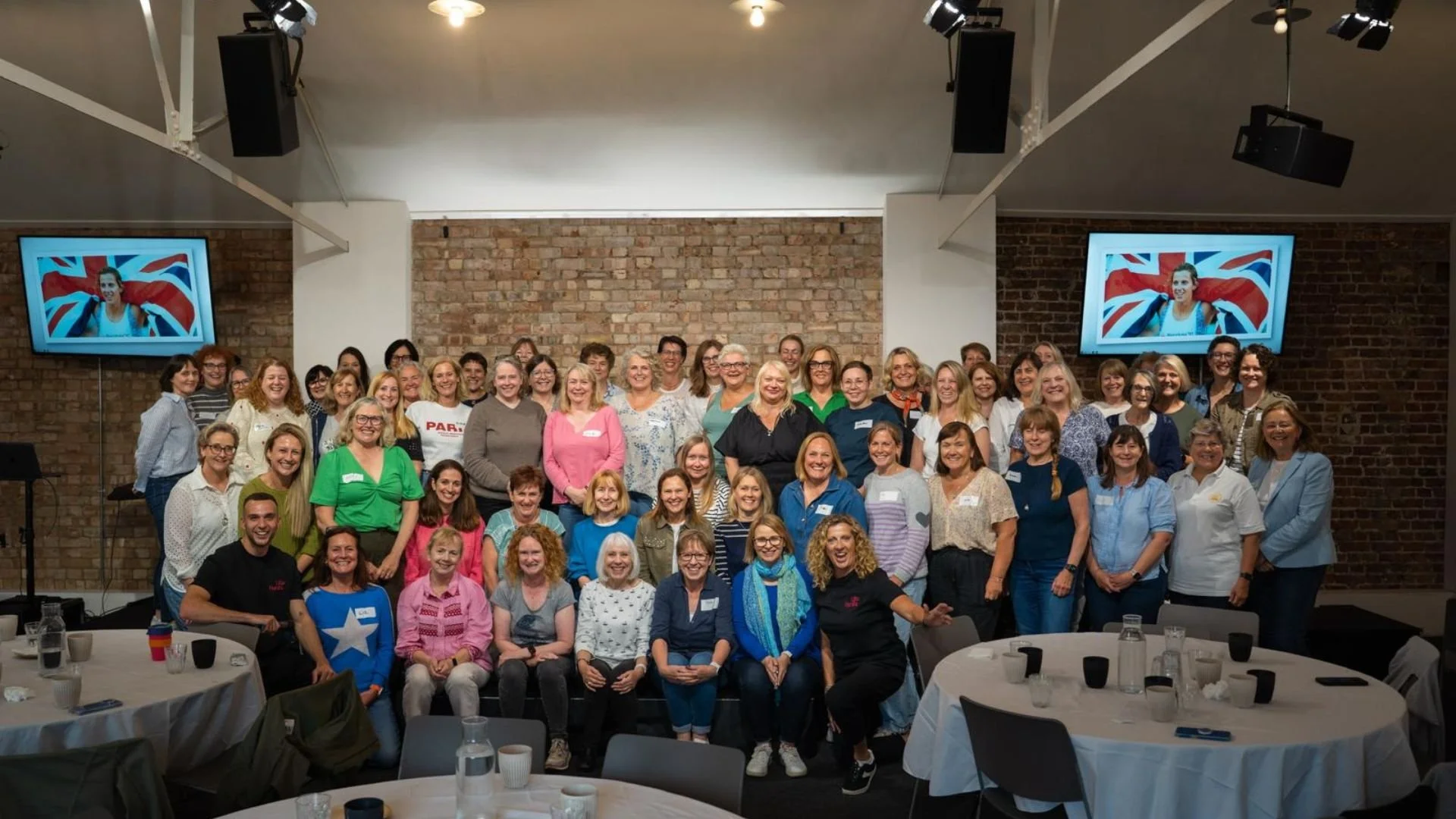 Group of smiling women and a few men gathered for a photo in a room with brick walls, with two monitors displaying a woman with a British flag background.