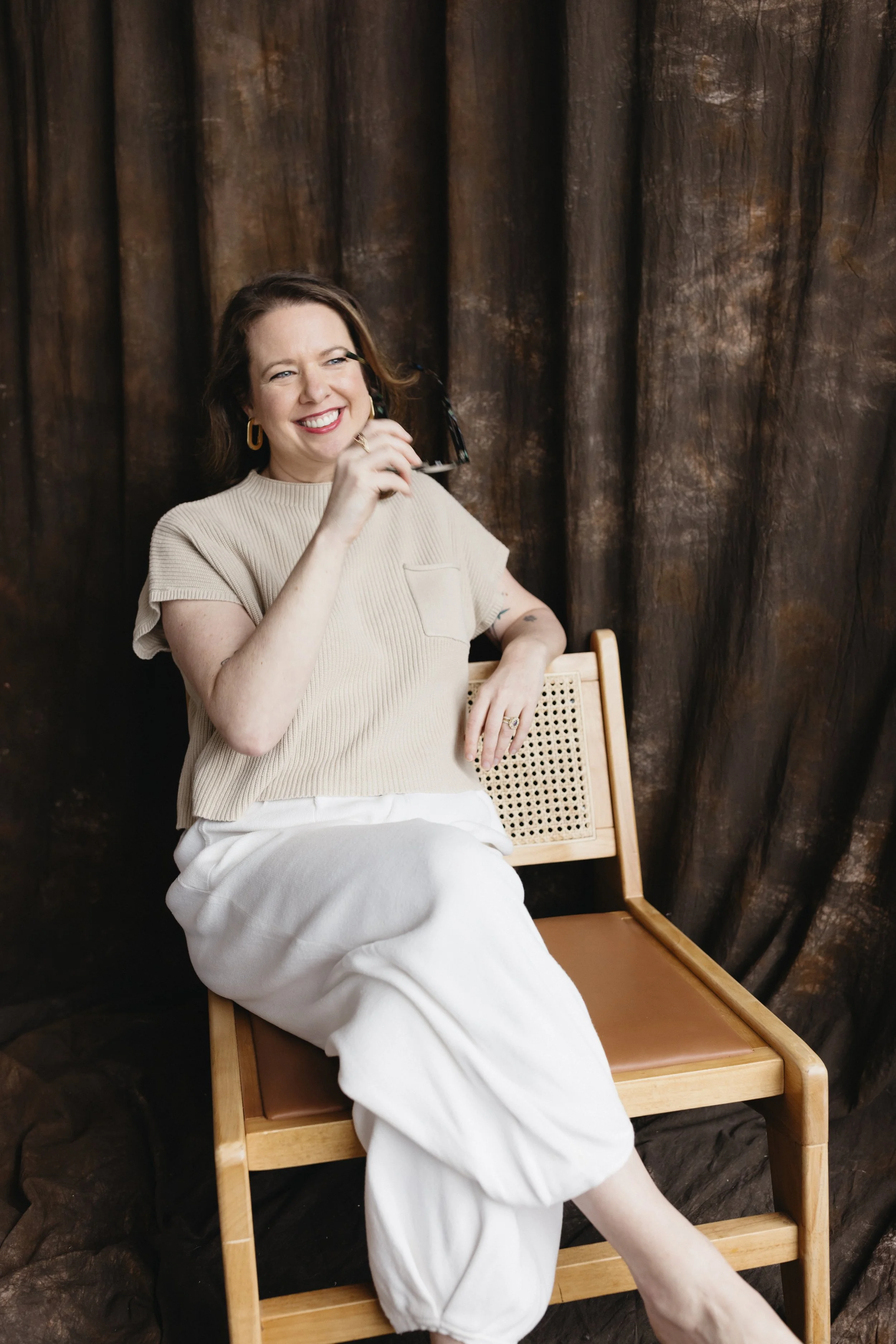 A woman sitting on a wooden chair, smiling, holding glasses, with a dark brown textured curtain background.