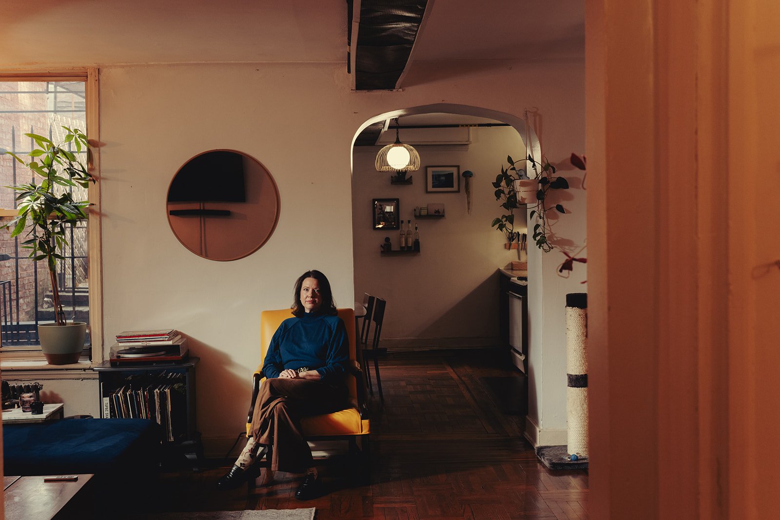 A woman sitting in a yellow chair in a cozy living room with a large window, plants, a coffee table, and wall decorations.