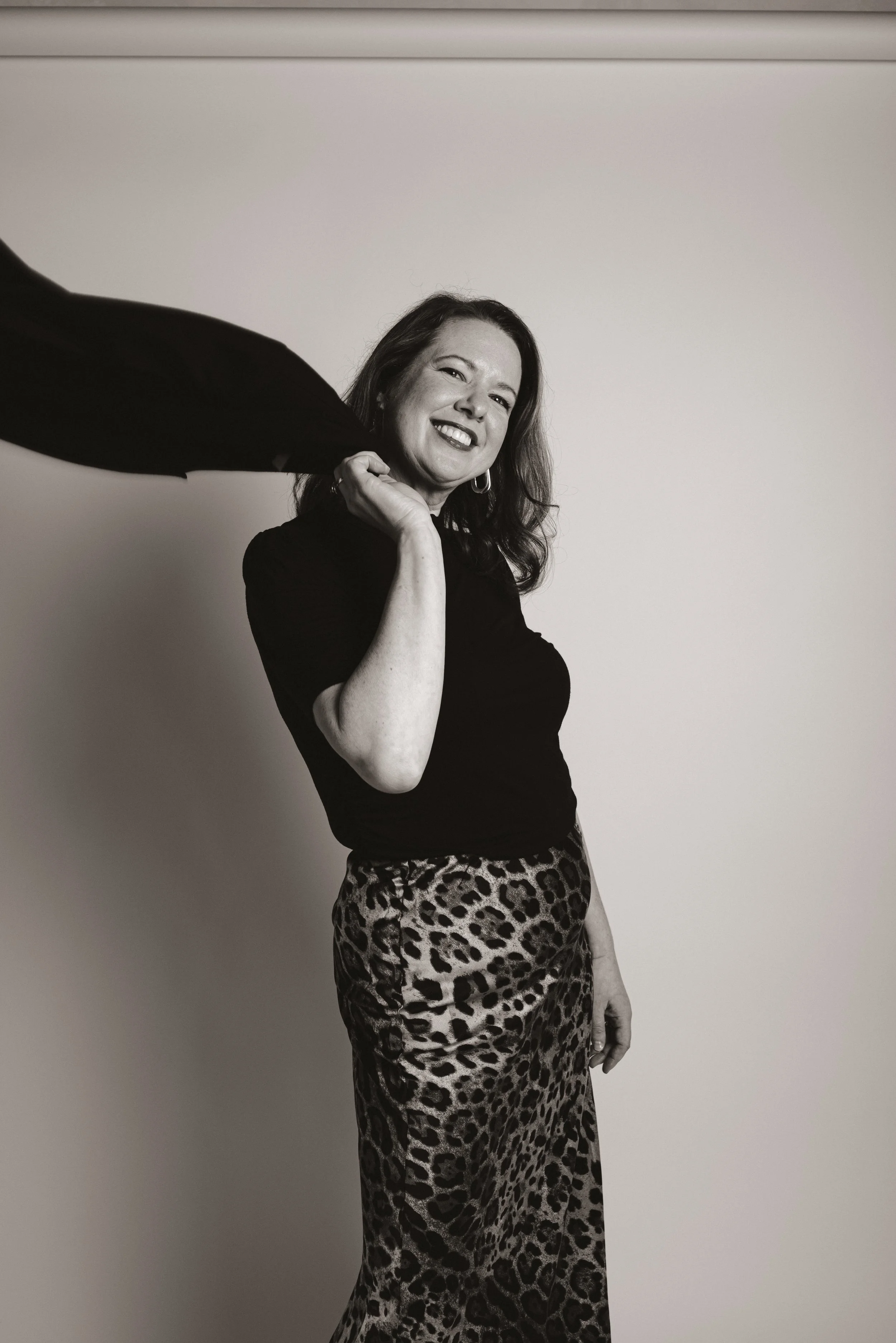 A woman smiling, holding a black fabric or scarf behind her head, wearing a black top and leopard print skirt, standing against a plain wall.