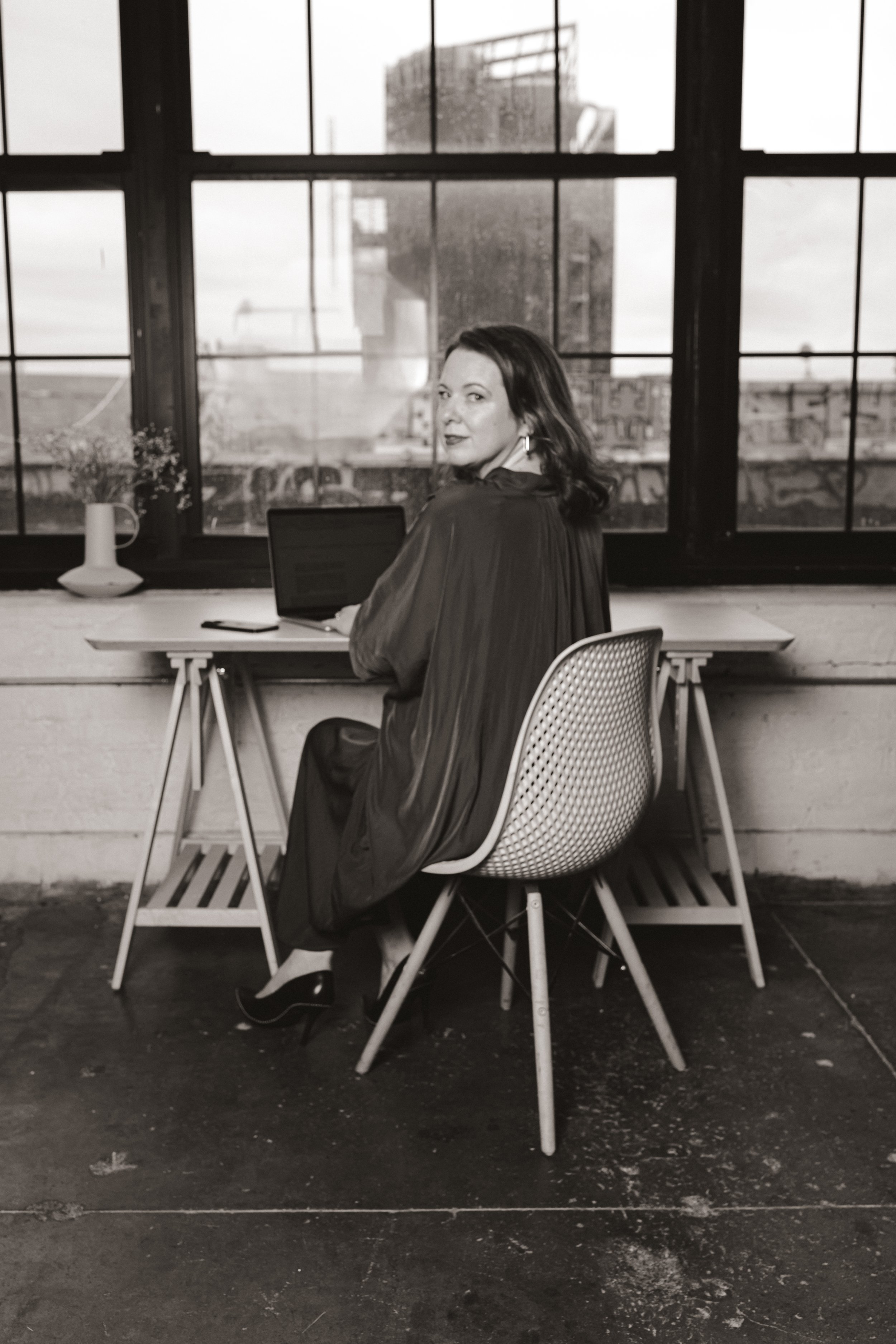Black and white photo of a woman sitting at a desk by a large window, working on a laptop, inside an industrial-style space.