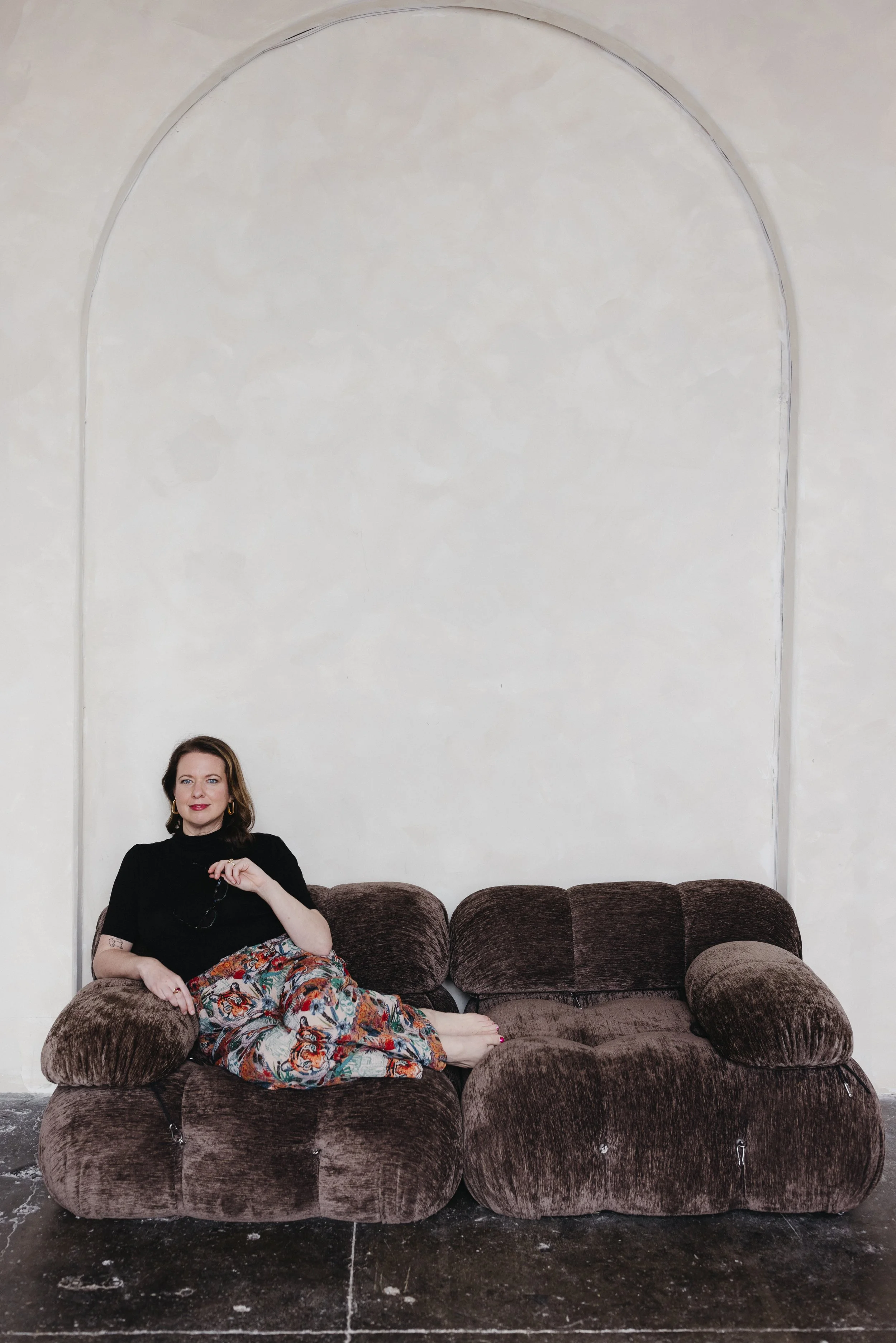 Woman sitting on a brown plush sofa in a minimalist room with a high arched wall and a concrete floor.