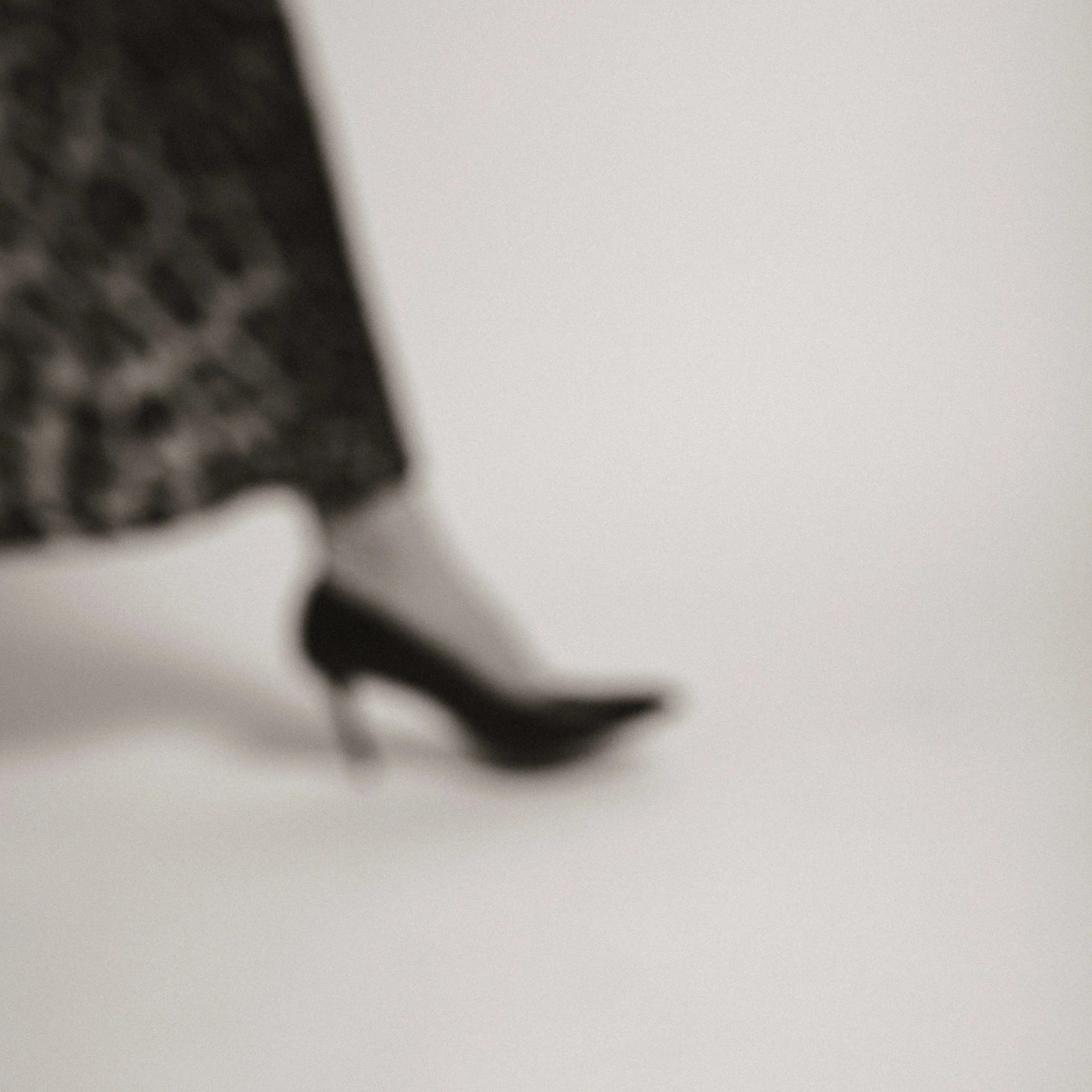 Close-up of a black high-heeled shoe worn with a patterned dress against a plain background.