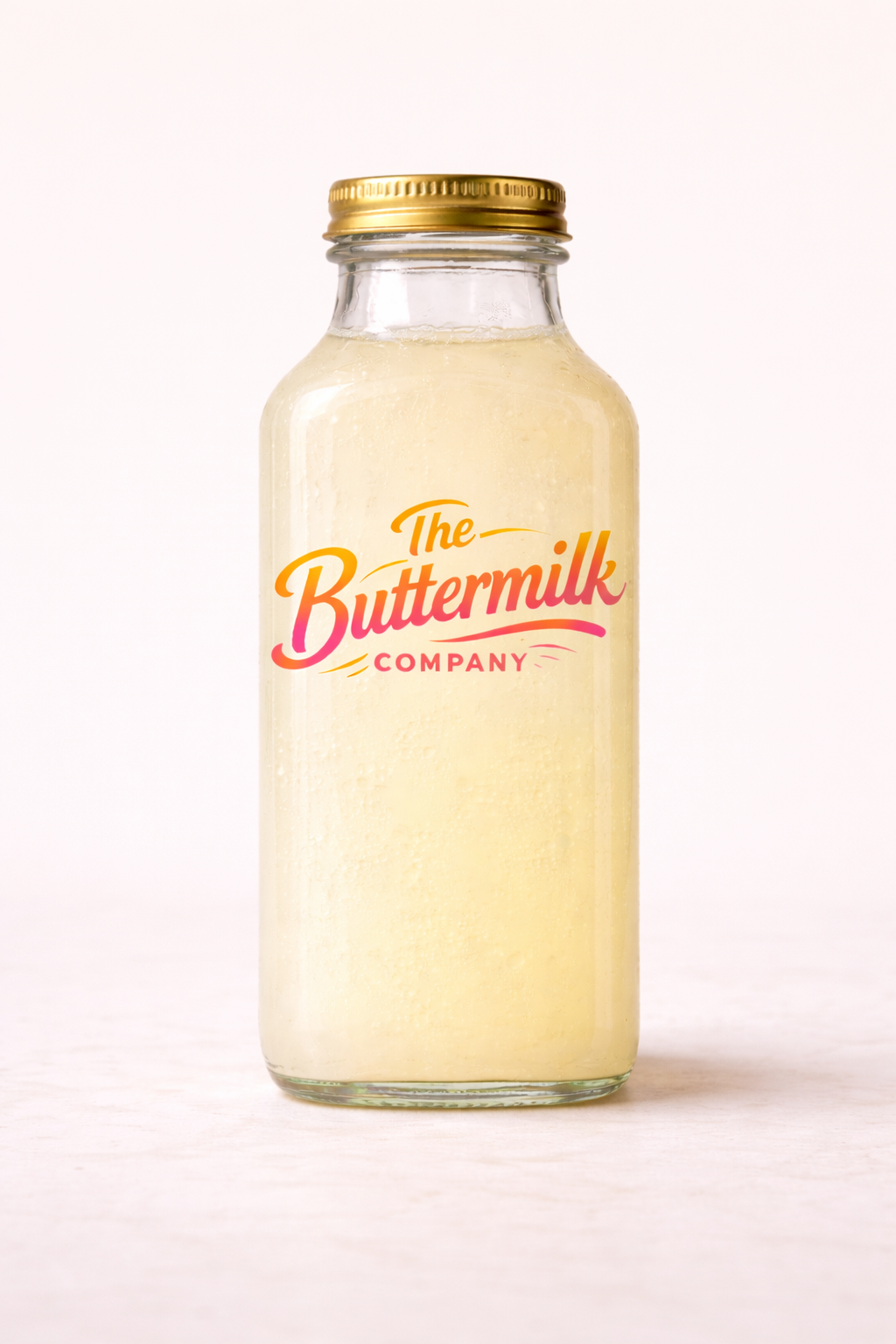 A glass bottle of buttermilk with a gold screw cap, labeled "The Buttermilk Company," against a plain white background.