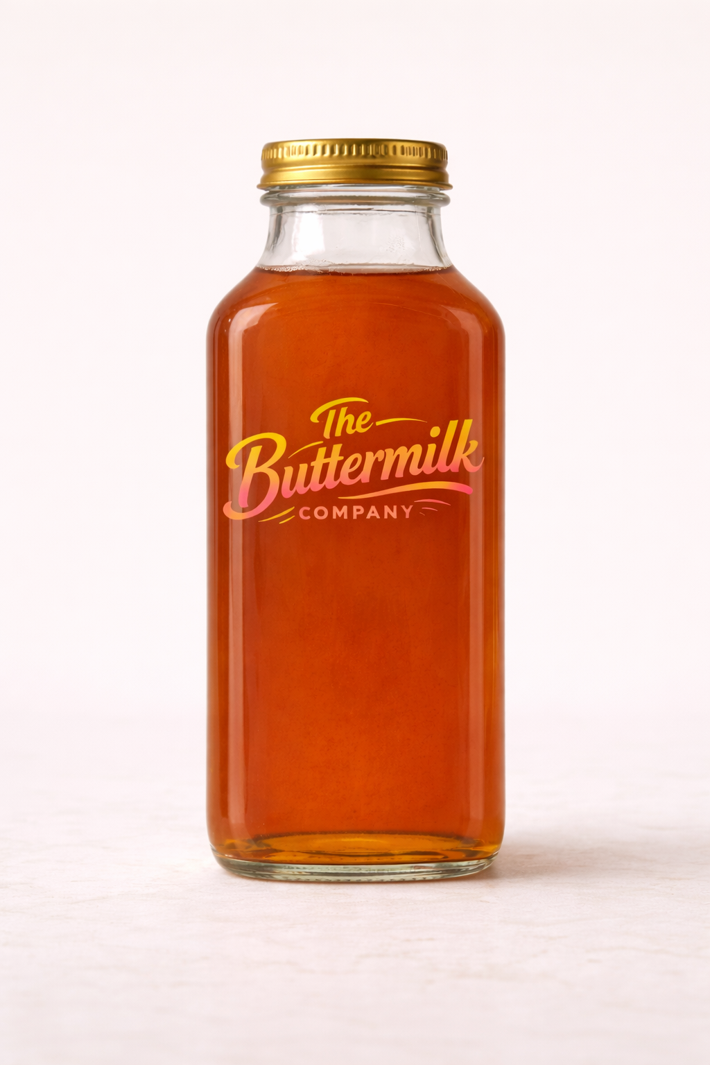 A glass jar filled with amber-colored liquid, labeled 'The Buttermilk Company' in pink and yellow font, with a gold lid, on a white background.