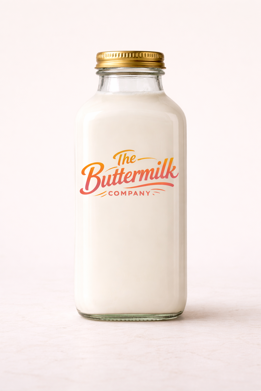 A glass bottle filled with milk with a gold lid and a logo reading 'The Buttermilk Company' in orange and pink lettering.