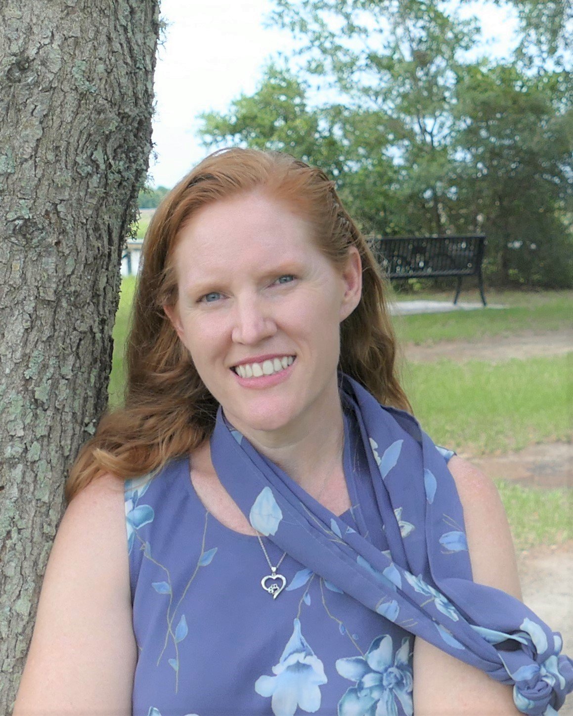 Author Dorothy McFalls sitting outdoors near a tree with a smile, wearing a blue floral dress and a heart-shaped necklace, with a park bench and green trees in the background.