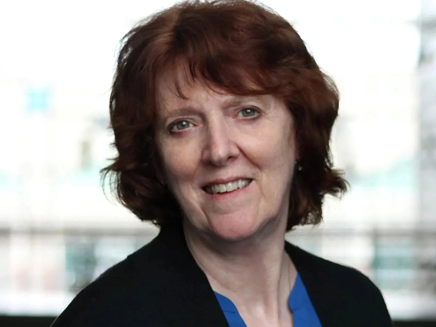 Author Robin Lee close up picture. She has short red hair, green eyes, and a friendly smile, wearing a black blazer and a blue top, standing in front of a blurred window background.