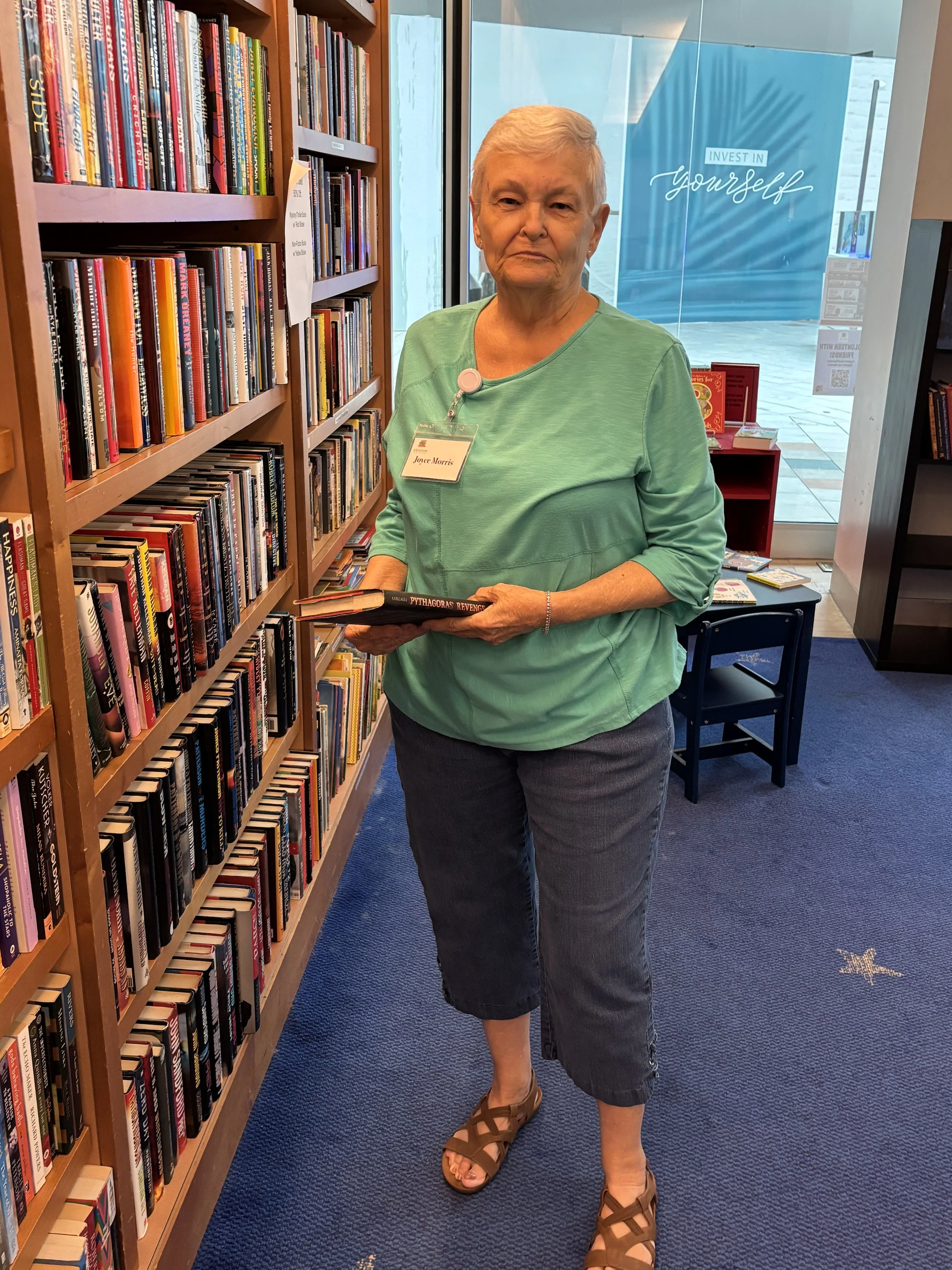 woman with short gray hair stands in a bookstore or library aisle holding a book. She is wearing a light green blouse, dark gray cropped pants, and brown sandals. Behind her is a large window with a blue sign that says 'Invest in Yourself', and the shelves next to her are filled with various books.
