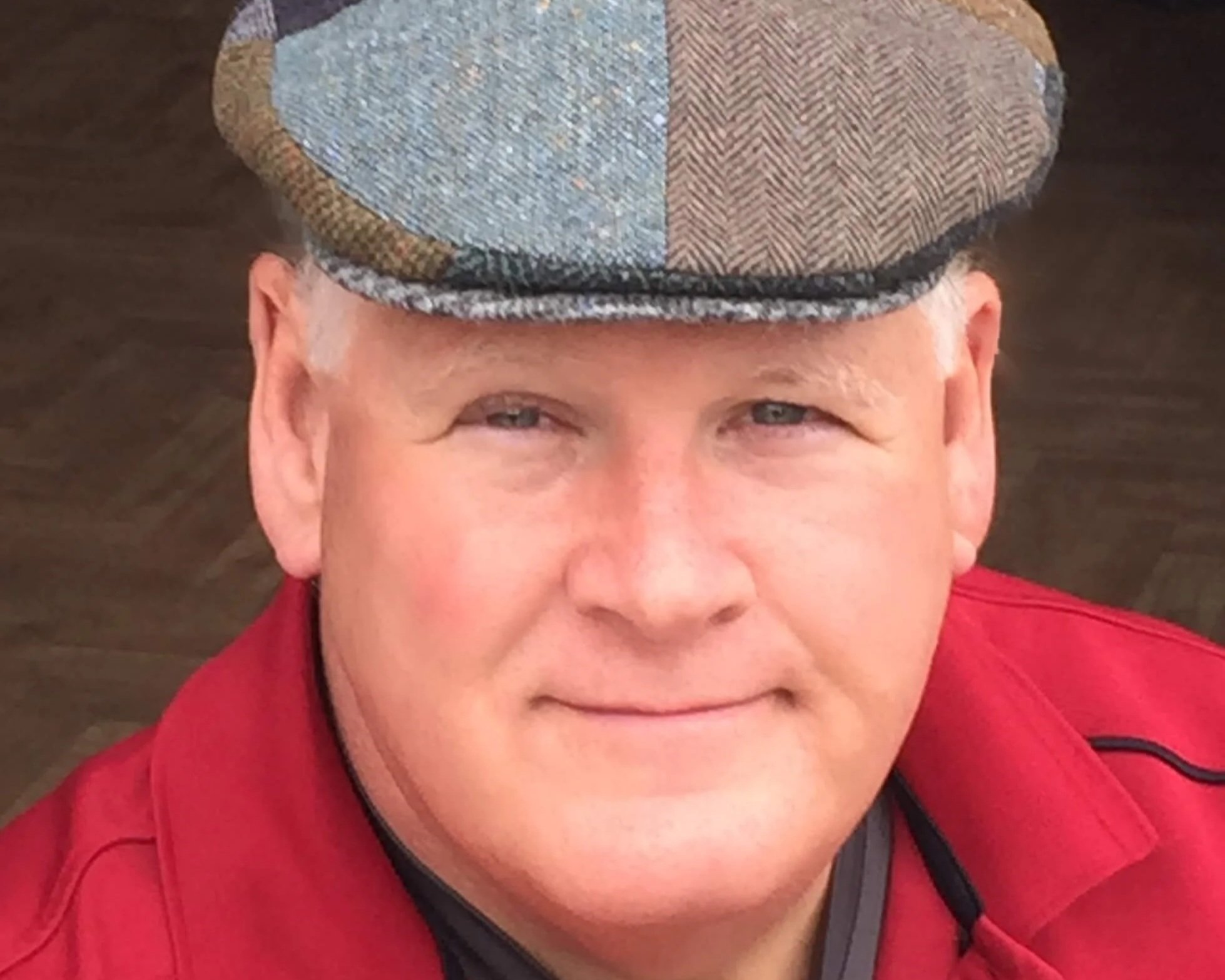 Author Roger Newman, a man wearing a plaid flat cap and a red jacket, smiling, with a wooden background.