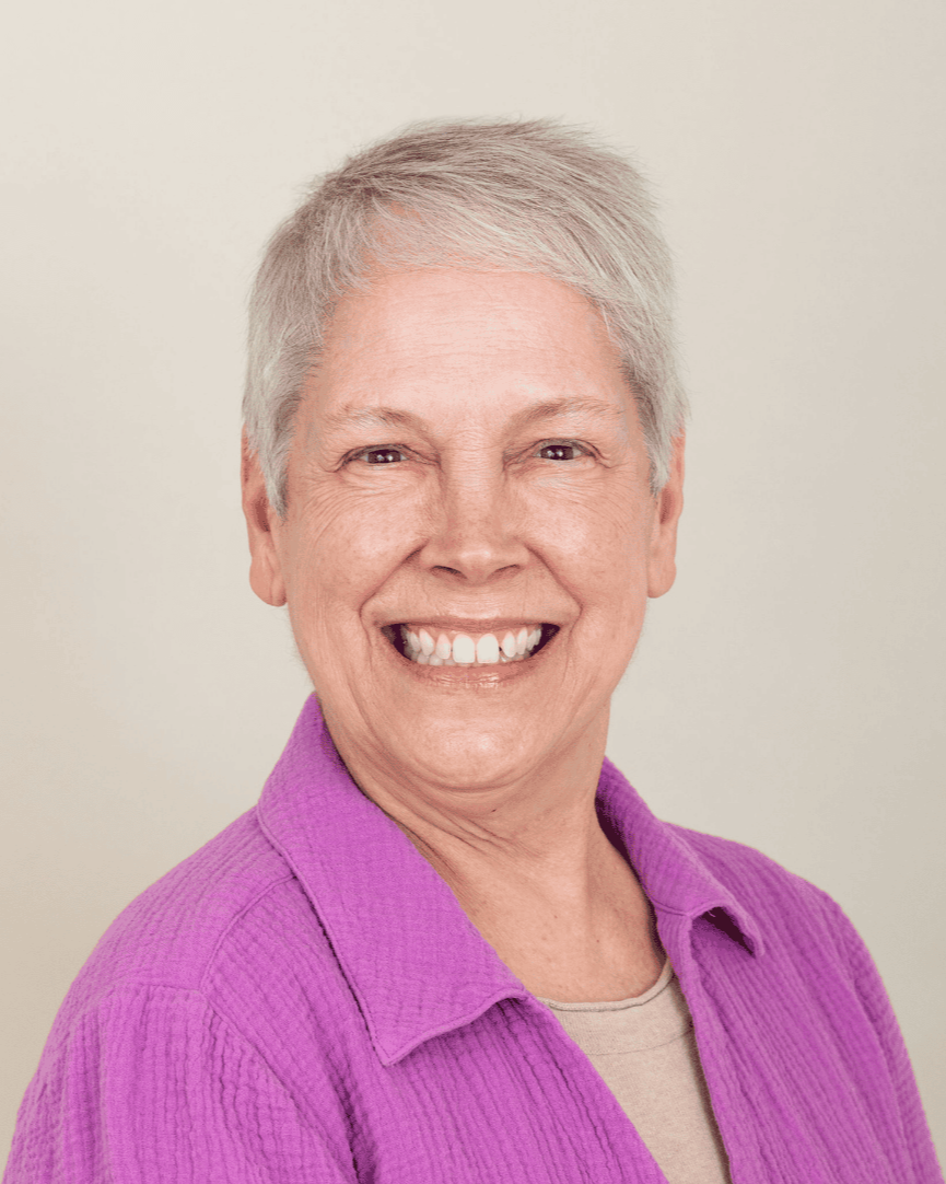 author Melissa Henderson - A smiling woman with short gray hair, wearing a blue shirt, standing indoors with a blurred building in the background.