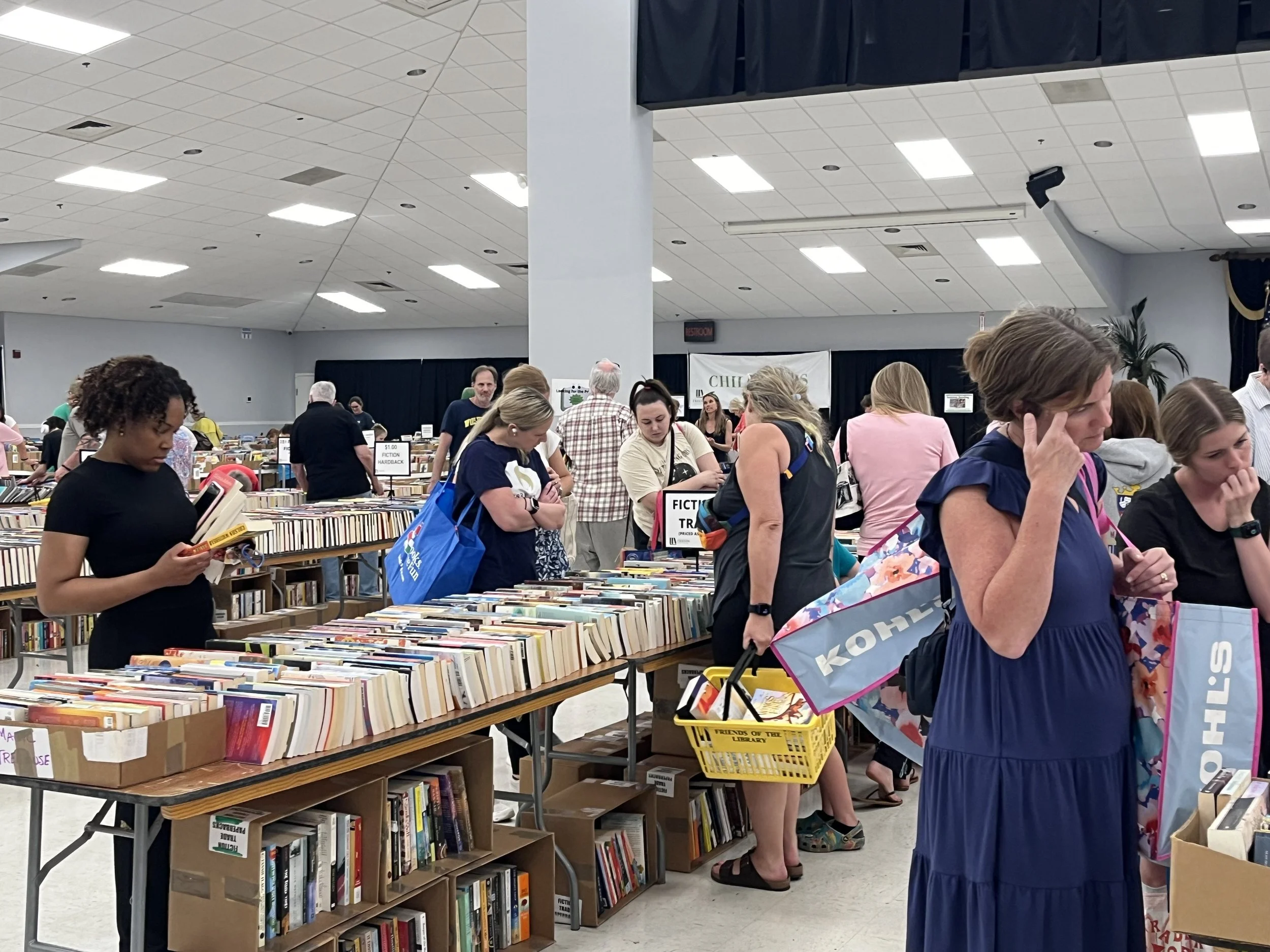 People browsing books at a book sale with tables filled with books in a large indoor space.