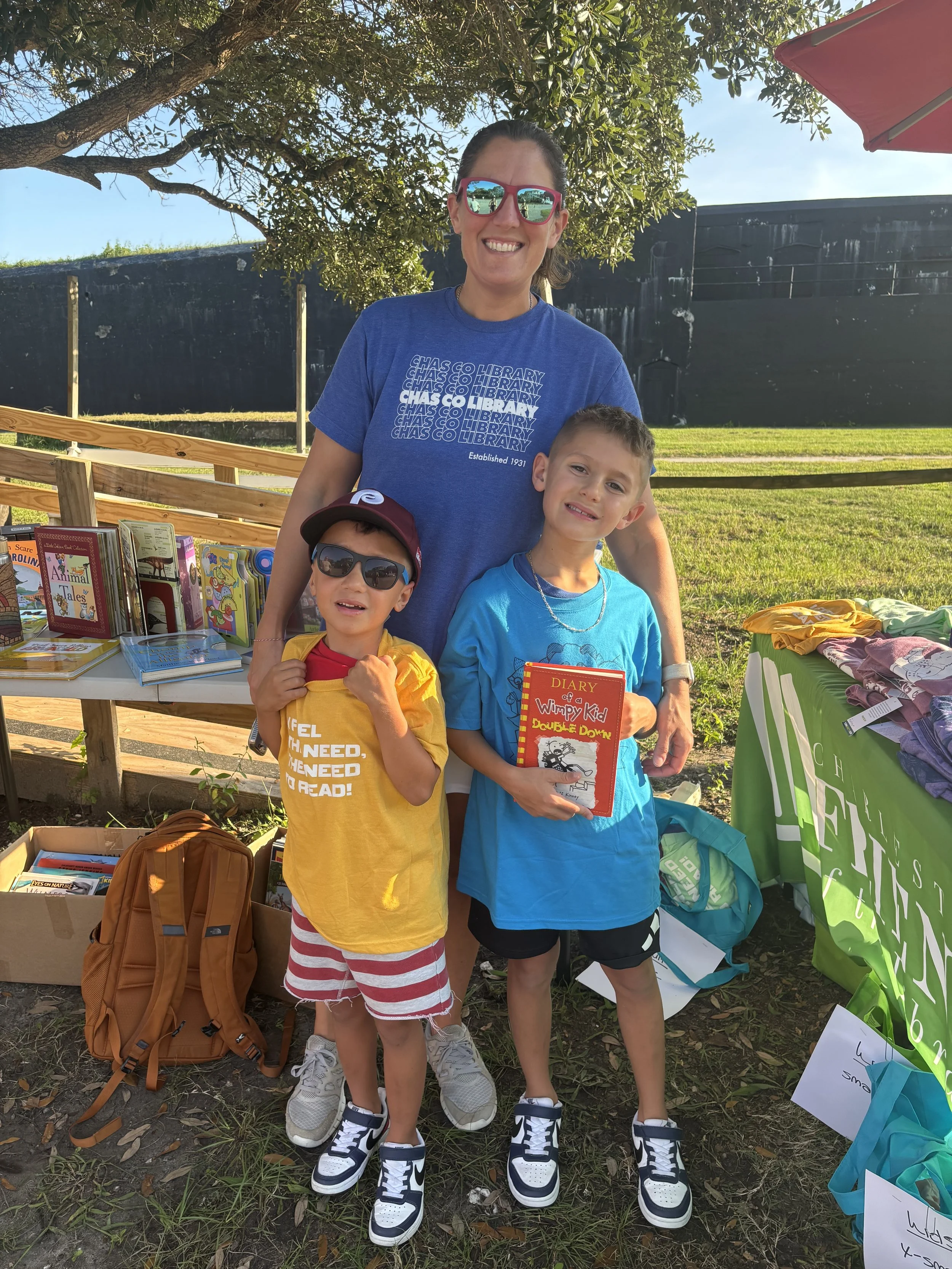 A woman and two young boys standing outdoors at a book sale table with books and clothes displayed. The woman is smiling, wearing sunglasses and a blue t-shirt. One boy, wearing sunglasses and a maroon cap, is holding a yellow shirt and is in front, while the other boy, holding a red book titled 'Diary of a Wimpy Kid', is dressed in a bright blue t-shirt. The scene has trees and a grassy area in the background.