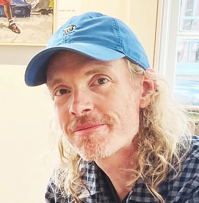 Author Tim Banks - A man with long curly blonde hair wearing a blue baseball cap and a checkered shirt, smiling indoors near a window and a wall with artwork.
