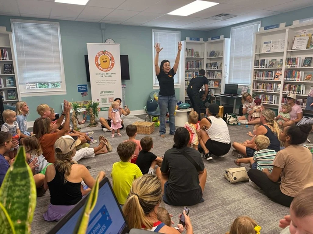 A woman leading a storytime or activity session with children and parents seated on the floor in a library. The woman has her arms raised, engaging the audience. The library has bookshelves filled with books, windows with blinds, and a banner in the background.