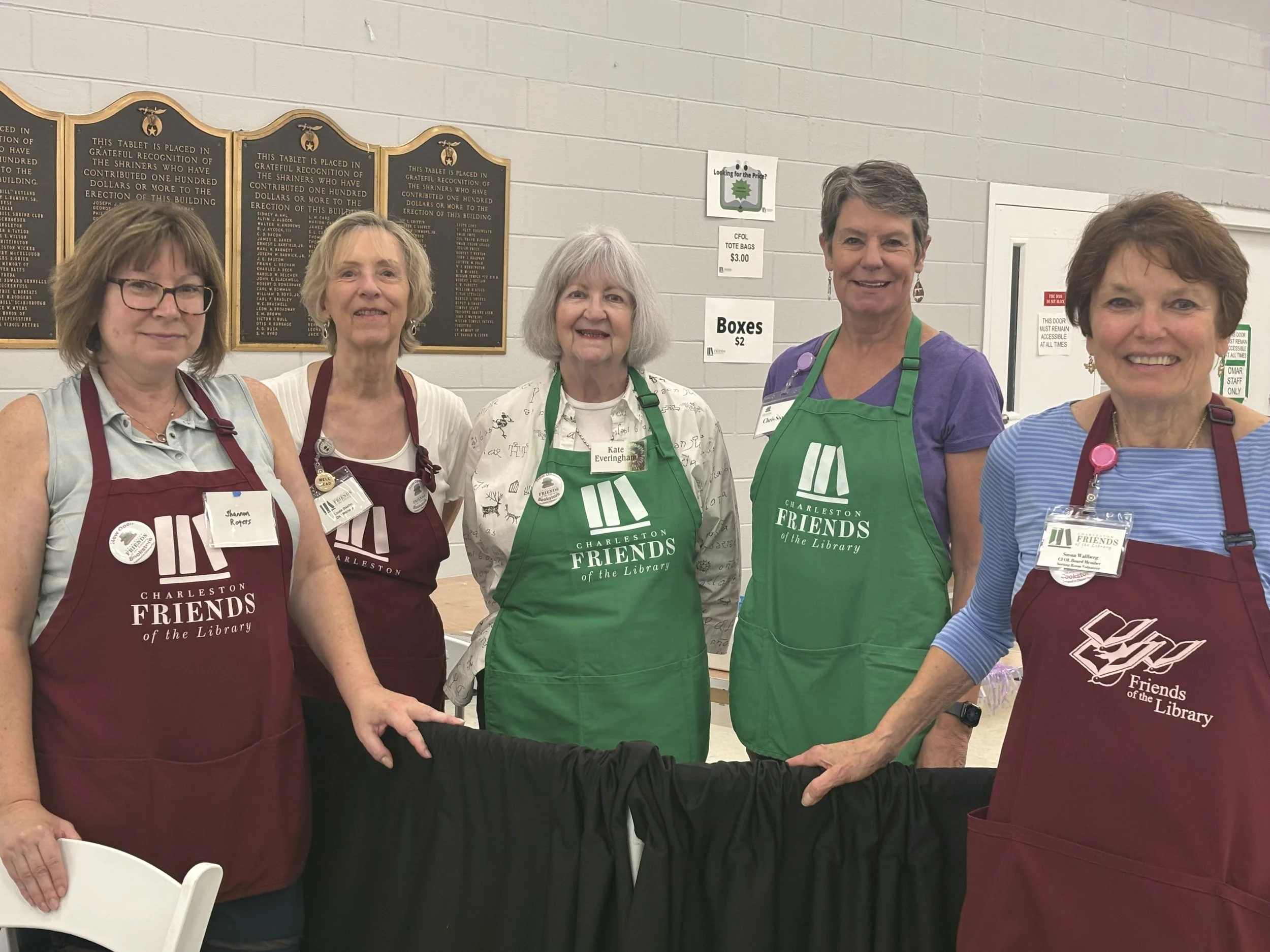 Group of five women wearing Charlestown Friends of the Library aprons, standing behind a table at a library event.