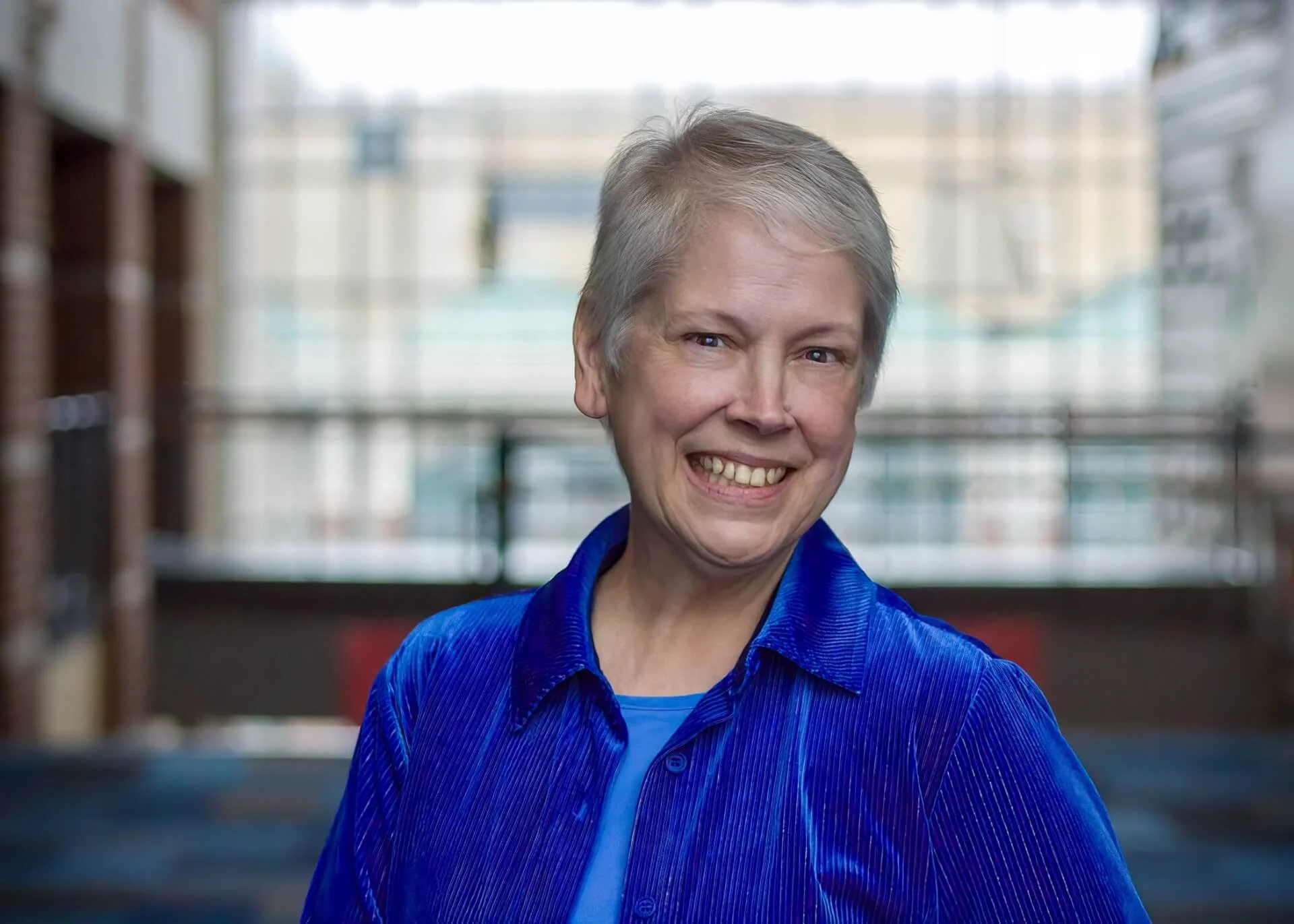 author Melissa Henderson - A smiling woman with short gray hair, wearing a blue shirt, standing indoors with a blurred building in the background.