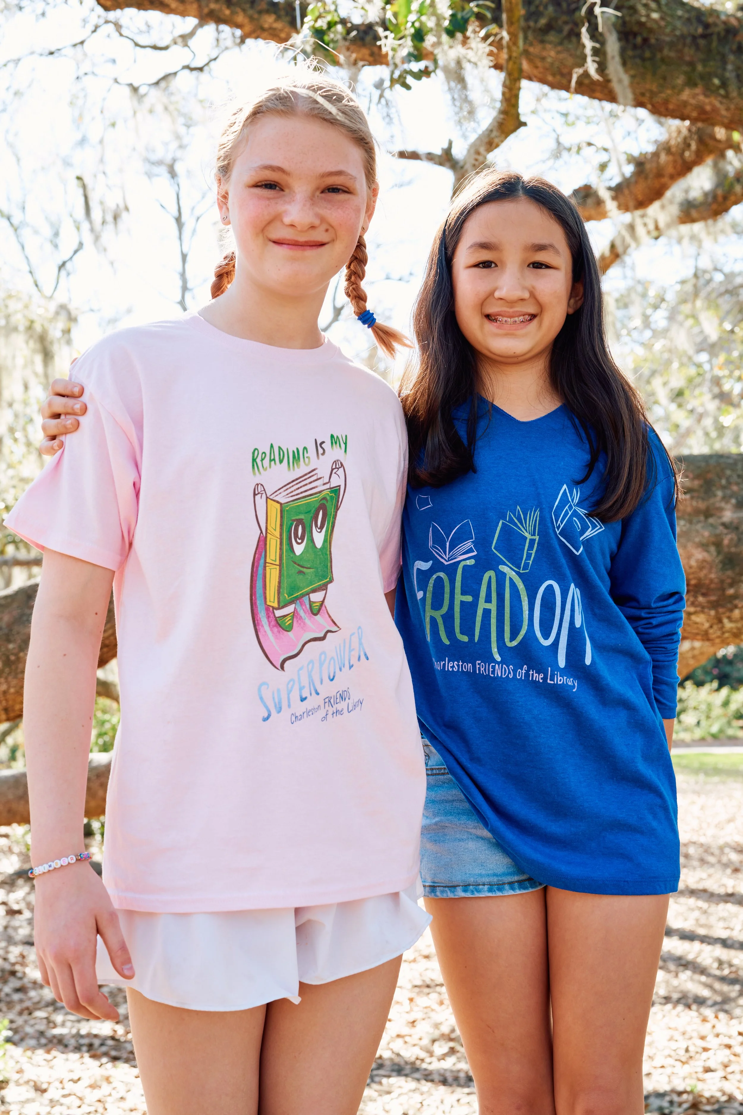Two young girls standing outdoors, smiling, with their arms around each other's shoulders. They are wearing colorful T-shirts with library-themed designs and text, under a tree.