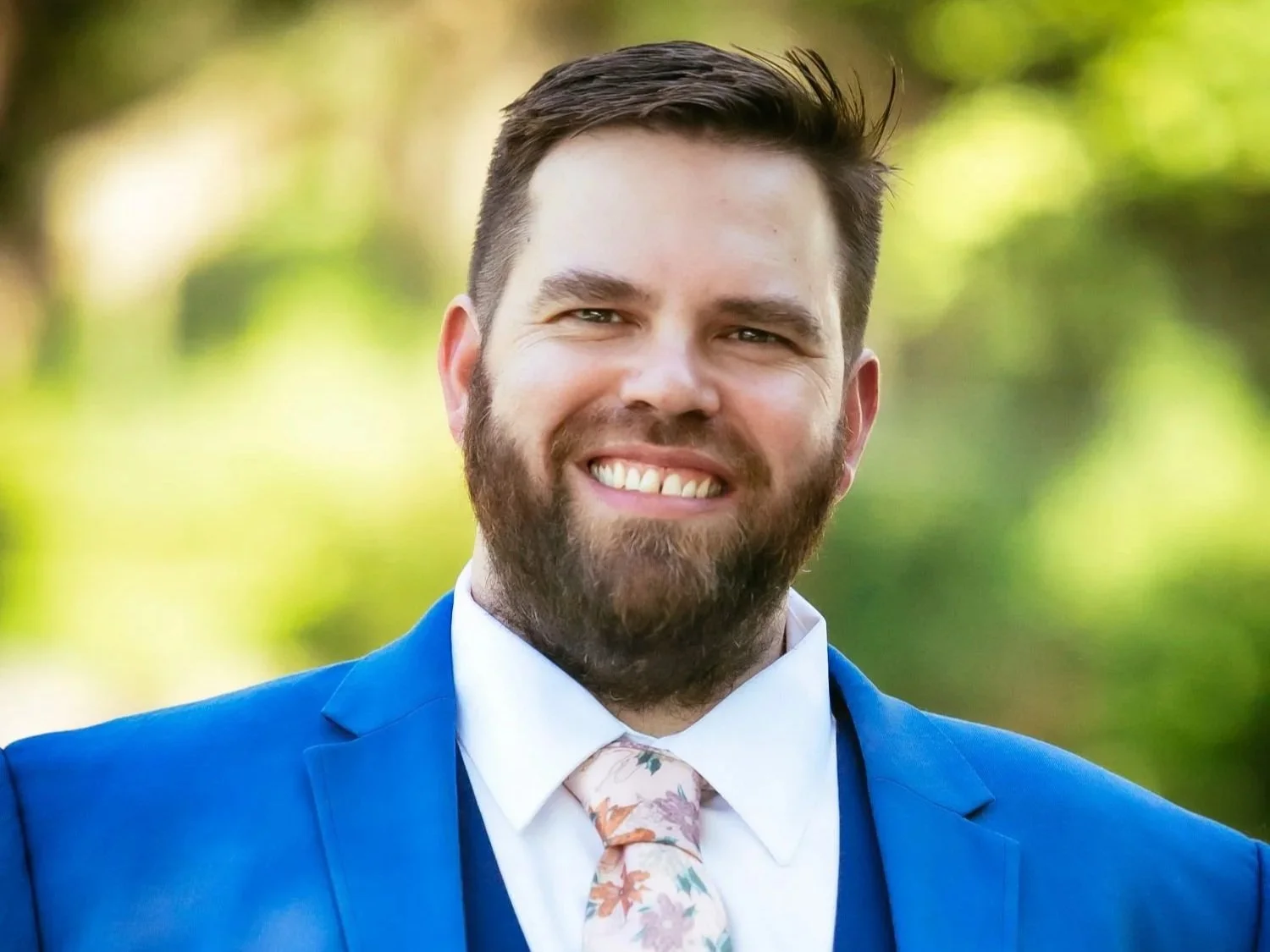 A smiling man, author Chris kinard, with brown hair and a beard, dressed in a blue suit, white shirt, and floral tie. Background is outdoors with greenery and blurred trees.