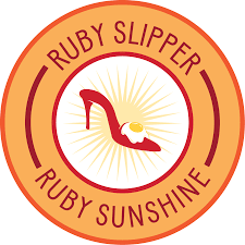 Logo for Ruby Slipper Ruby Sunshine featuring a red high-heeled slipper with a yellow sunray background inside a circular badge.