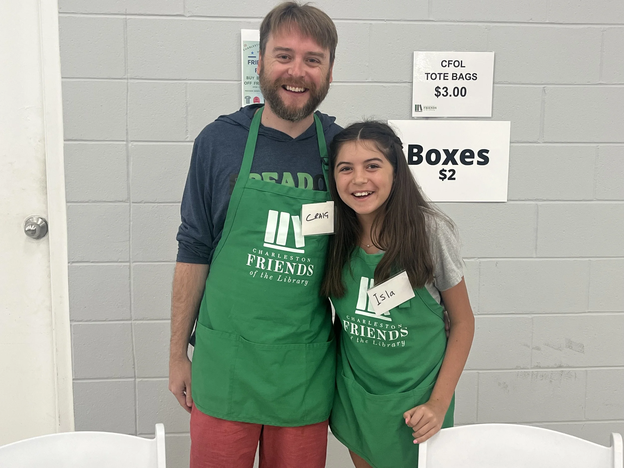 A man and girl wearing green aprons with Charleston Friends of the Library logos smiling indoors.
