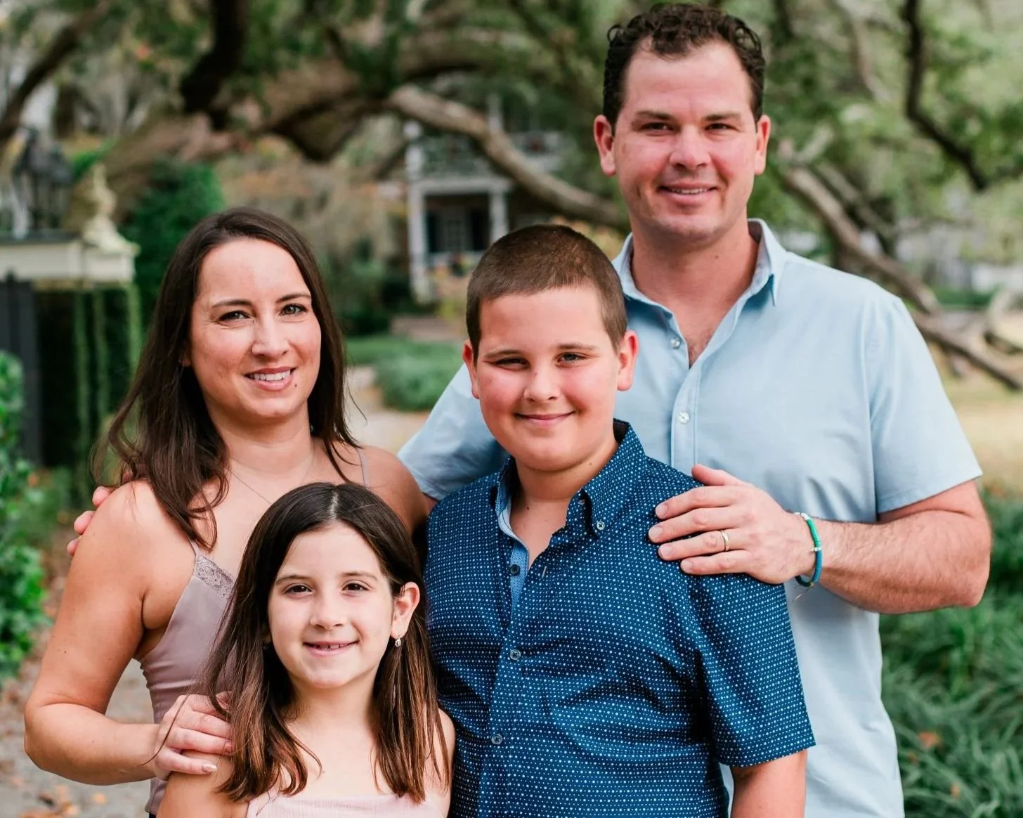The Yagecic family of five outdoors, smiling at the camera. The family includes a man, a woman, a teenage girl, and two boys, all standing in front of trees and a house.
