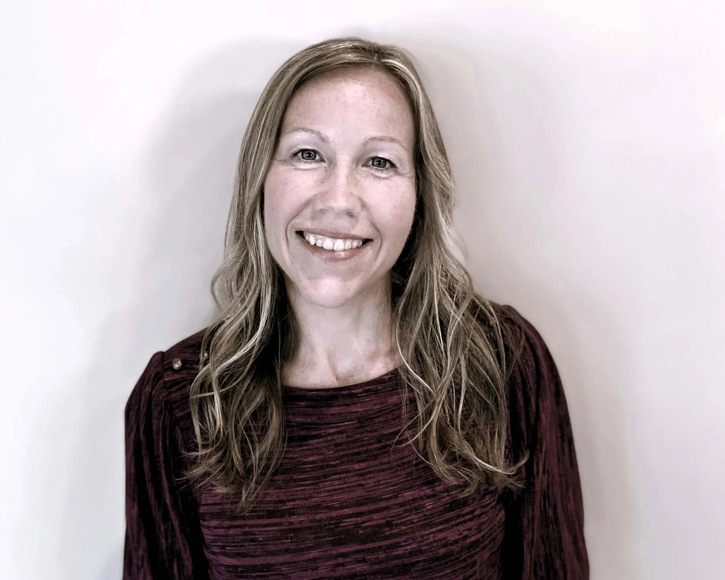 Author Abagail Summers, smiling with long wavy blonde hair wearing a dark red long-sleeve shirt standing against a plain white wall.