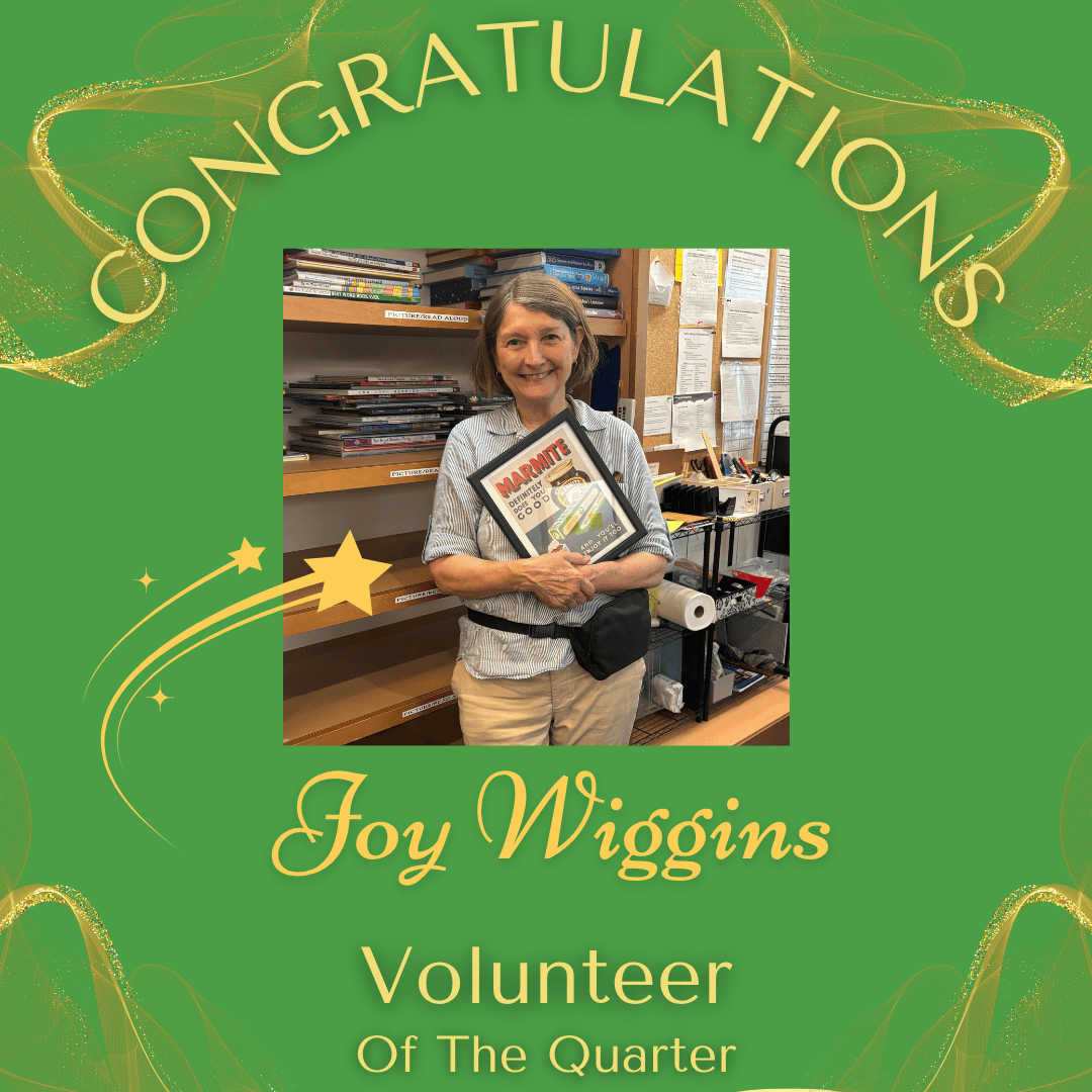 A woman smiling and holding a framed award that says 'Marmite.' She is standing inside an office or supply room with shelves of papers and supplies in the background. The image is part of a congratulatory graphic with a green background, golden stars, and the words 'Congratulations Joy Wiggins Volunteer Of The Quarter' in yellow and green text.