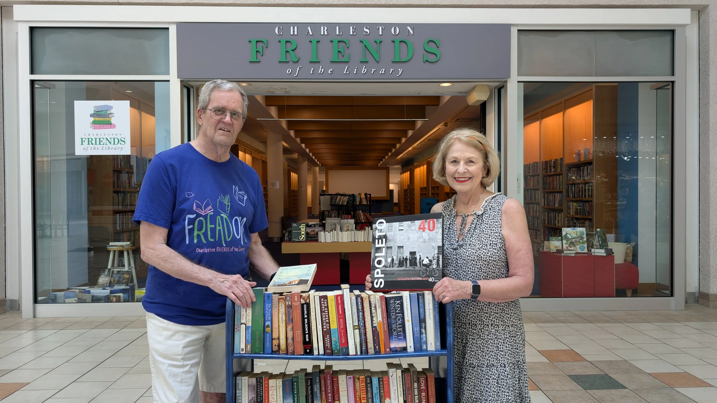 A man and woman standing in front of the Charleston Friends of the Library bookstore, holding novels and a book with a black and white cover titled "Spoleto 40." The woman has a basket of books beside her.