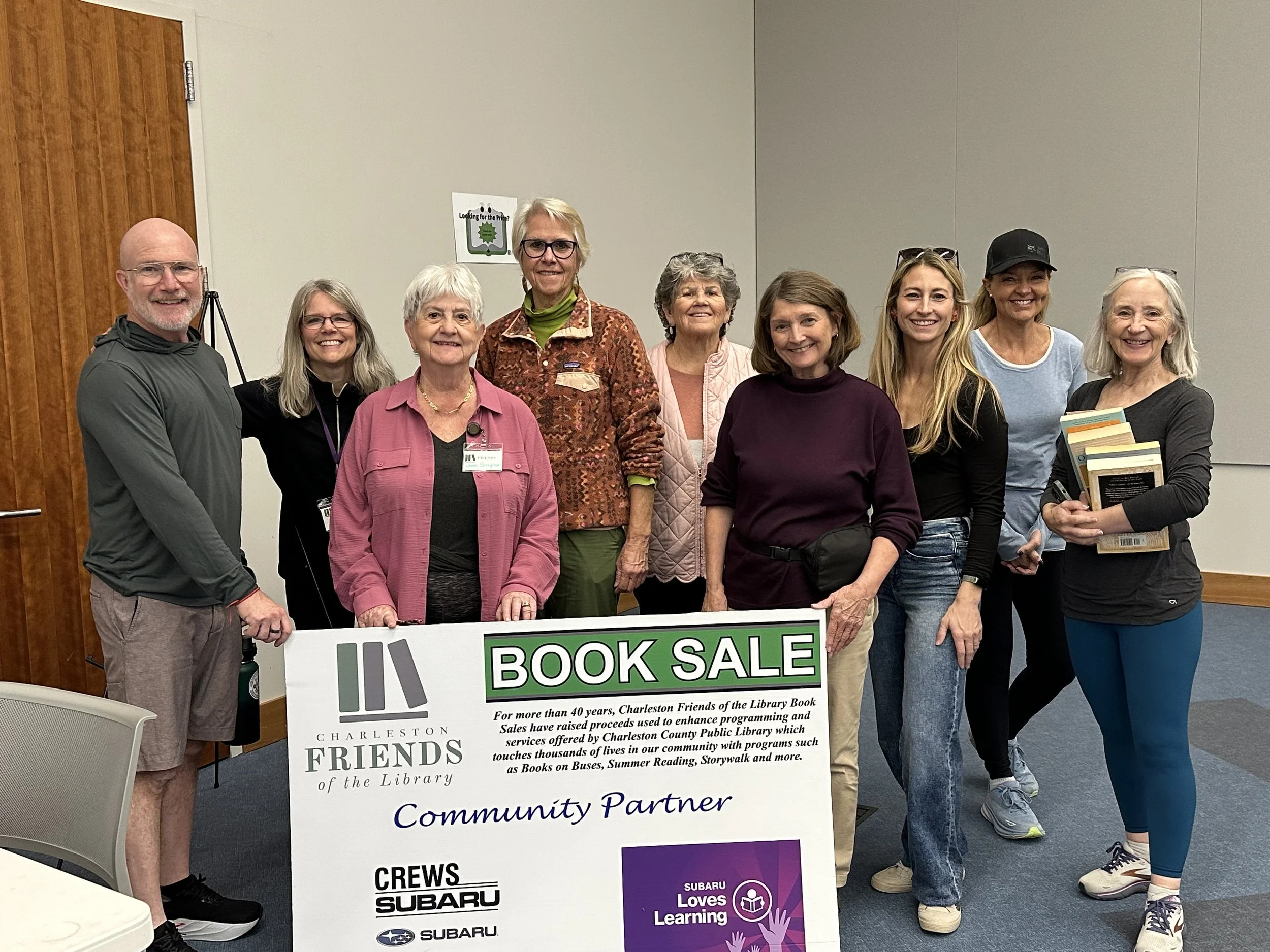 Group of ten smiling people standing behind a large sign that reads 'Book Sale Community Partner,' in a room with beige walls and wooden door.