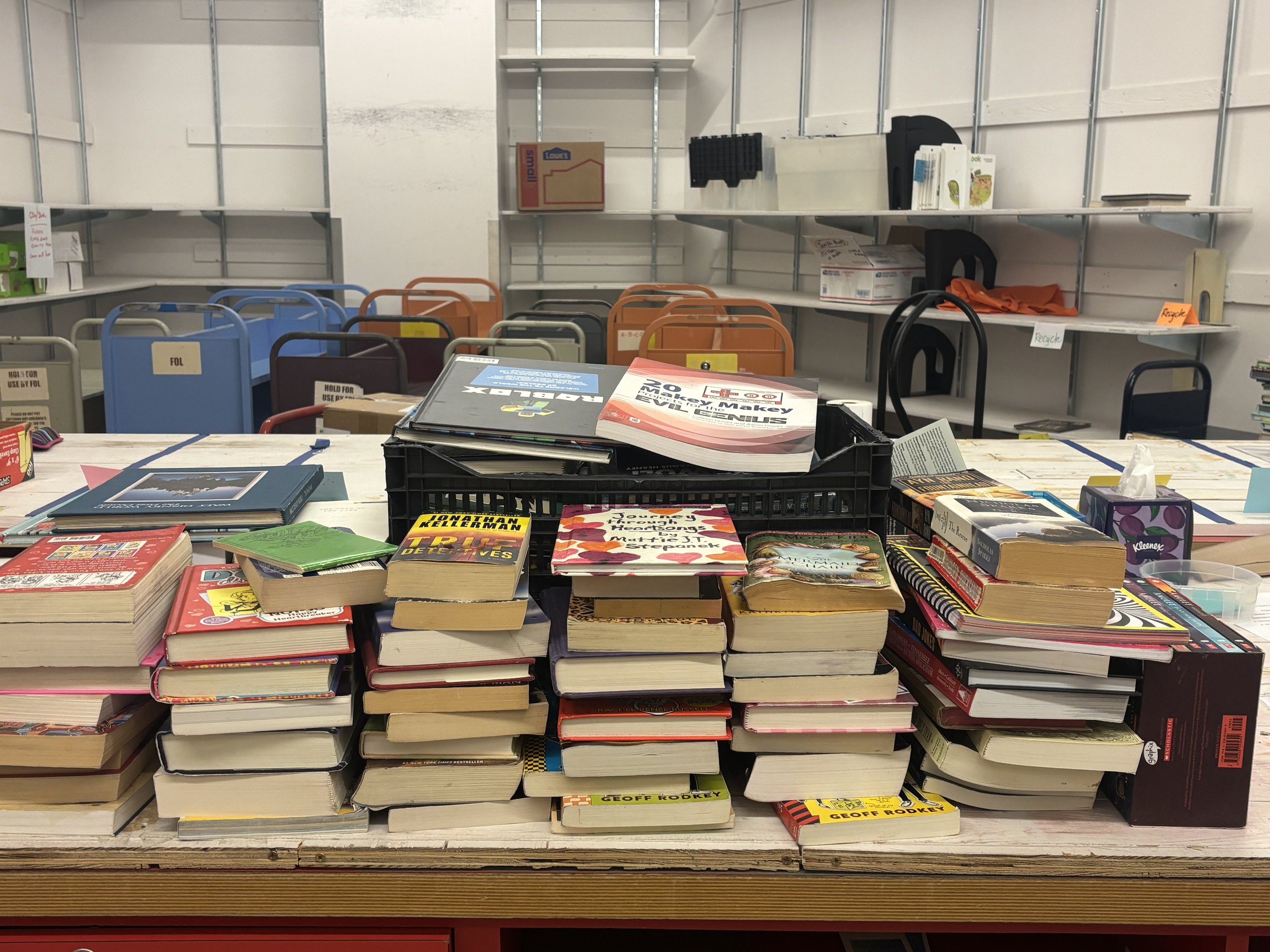 Table with stacks of books and magazines, with some papers and a box on top, in a storage or stockroom with chairs and shelves in the background.