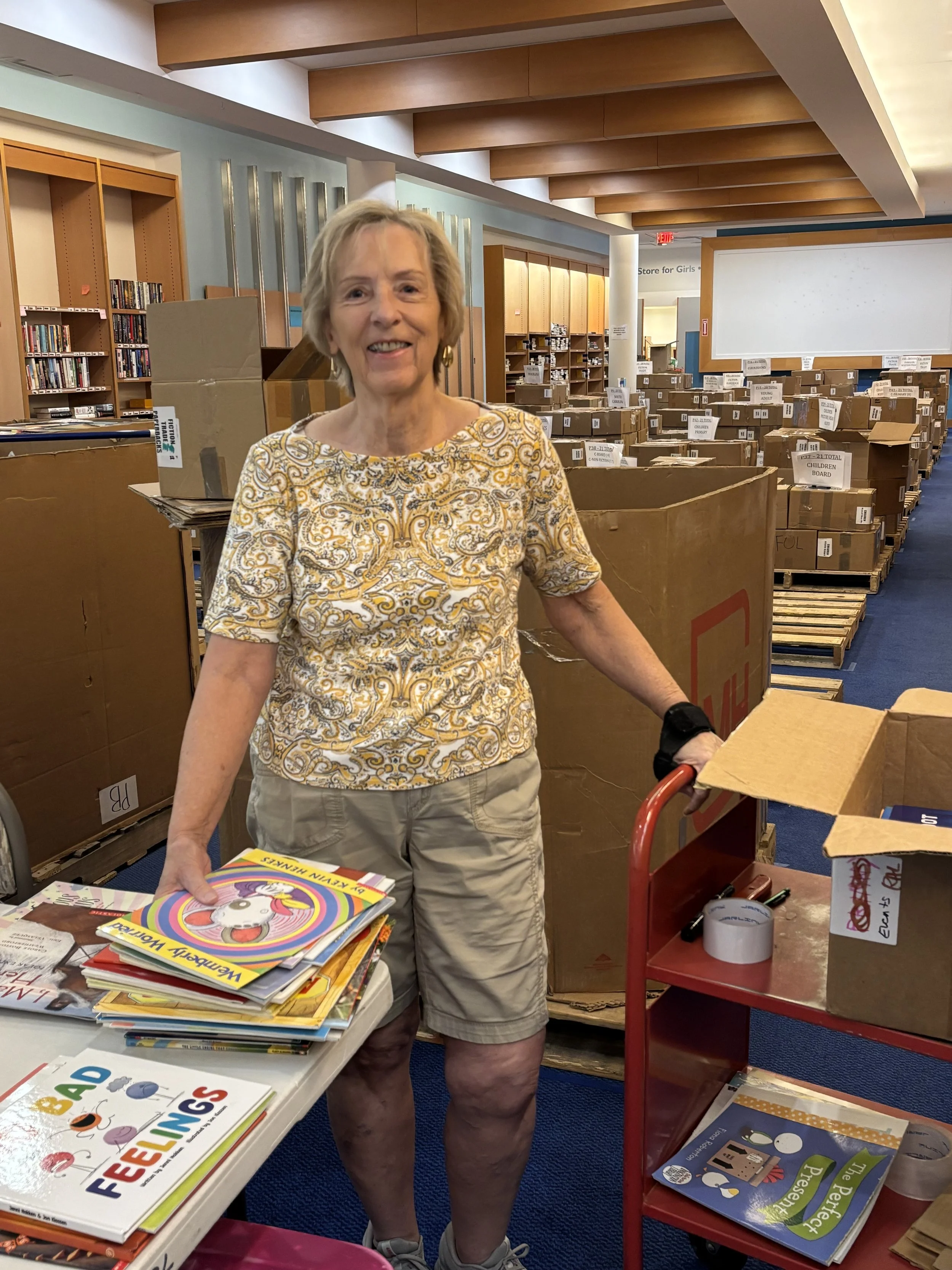 A smiling woman standing in the Charleston Friends of the Library bookstore at the Citadel Mall surrounded by cardboard boxes filled with books and magazines, next to a table with colorful children's books.